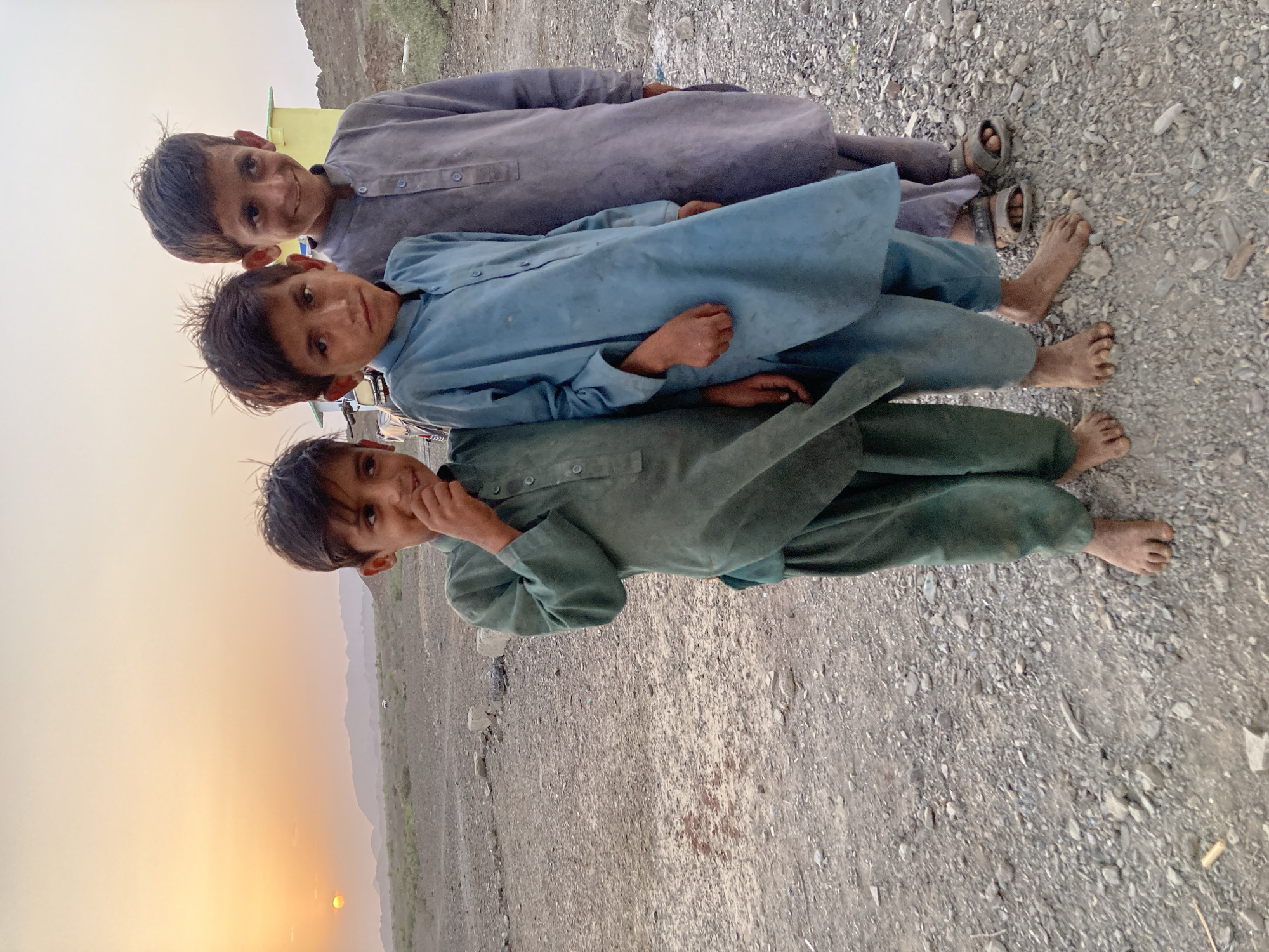 Three young boys stand close together in a vertical row on a gravelly surface. The top boy wears a gray outfit and sandals. The middle boy is in blue, and the bottom boy wears green. All three are barefoot. The background shows a landscape with a sunset. The location is Wayara, Pakistan.