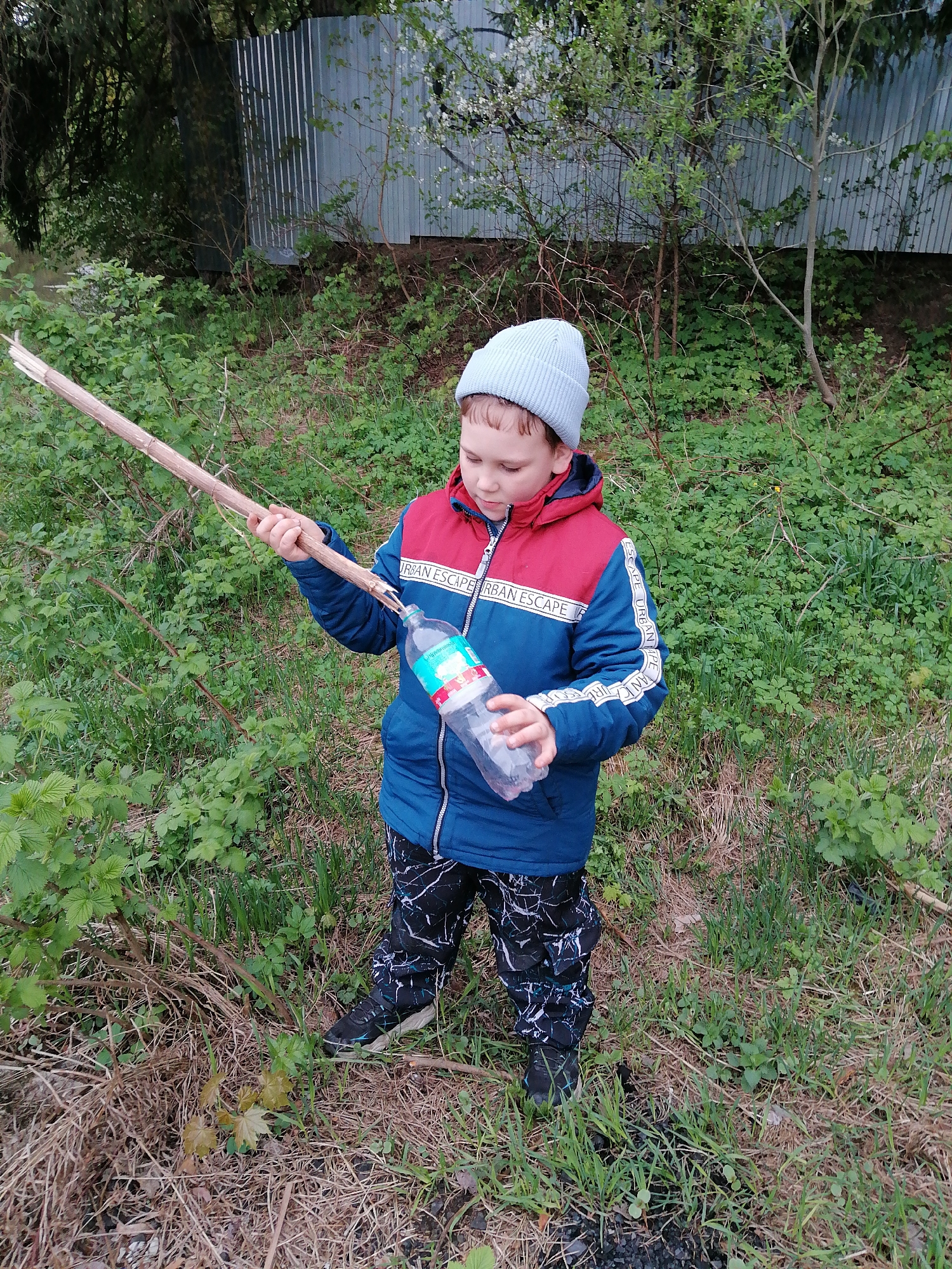 A young boy stands in a grassy area, dressed in a blue and red jacket, a light blue beanie, and patterned pants. He holds a long wooden stick in one hand and a clear plastic bottle in the other. The background consists of foliage and a corrugated metal fence. The scene appears to be outdoors in a naturalized area, possibly on the edge of a city or town. The location is the city of Unknown, Unknown.