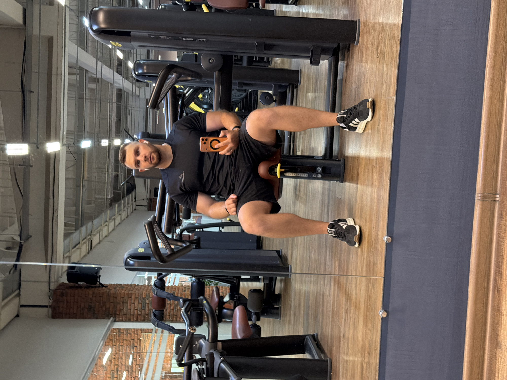 A man is sitting on a weight machine in a gym, holding his phone. He is wearing a black t-shirt and black shorts with black and white athletic shoes. The gym has wooden floors and exposed brick walls. The machines are black with brown padding. The lighting suggests it is daytime. The man has a neutral expression.