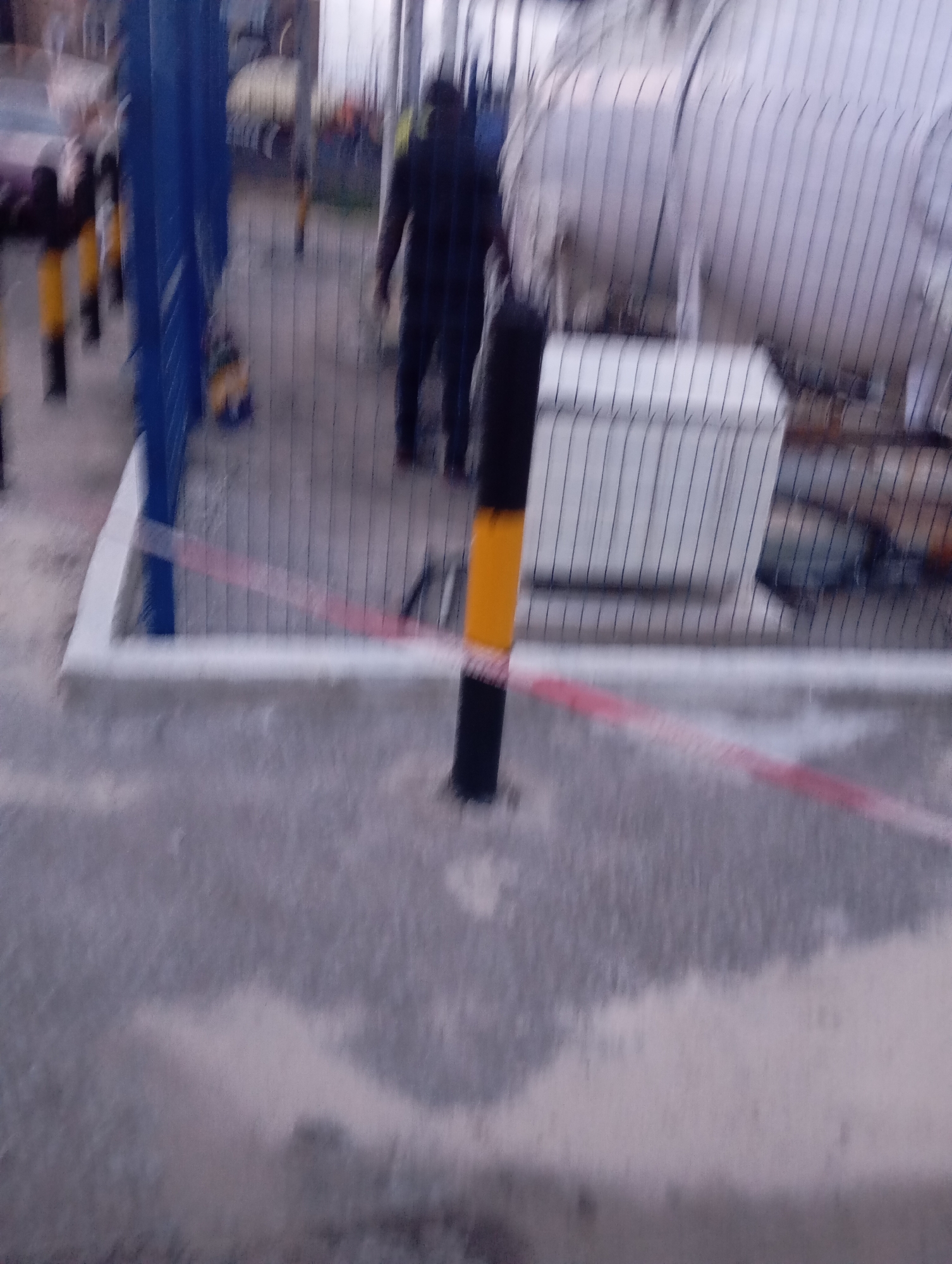 The image depicts an outdoor industrial area, likely within a facility in Port Harcourt, Nigeria, given the context. In the foreground, a grey, textured ground surface is visible, interspersed with patches of lighter colored material, possibly sand or dust. A black and yellow striped bollard stands prominently in the mid-ground, partially obscuring a red and white caution tape that stretches diagonally across the frame. Beyond the bollard, a chain-link fence separates the foreground from the background. Through the fence, a large, white cylindrical tank is visible, suggesting the presence of an industrial or storage facility. To the left of the tank, a white, rectangular structure with a ribbed surface is present, possibly a control box or storage unit. In the far background, a person, appearing to be male and wearing dark clothing, is walking away from the camera. Other figures and objects are blurred and indistinct due to the image's low resolution and motion blur. The overall impression is that of an active industrial site.