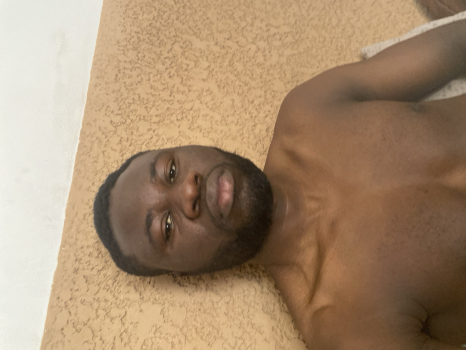 This image depicts an adult male, identified as being in Udu, Nigeria. The subject, of dark complexion, has short dark hair and a dark, trimmed beard and mustache. He is shirtless, with his bare chest and shoulders visible, facing directly towards the viewer with a neutral expression and slightly pursed lips. His gaze is directed forward. The background consists of a light brown or tan wall featuring a stucco-like texture, which meets a plain white ceiling in the upper portion of the frame. The scene is illuminated by uniform lighting, indicating an indoor environment.