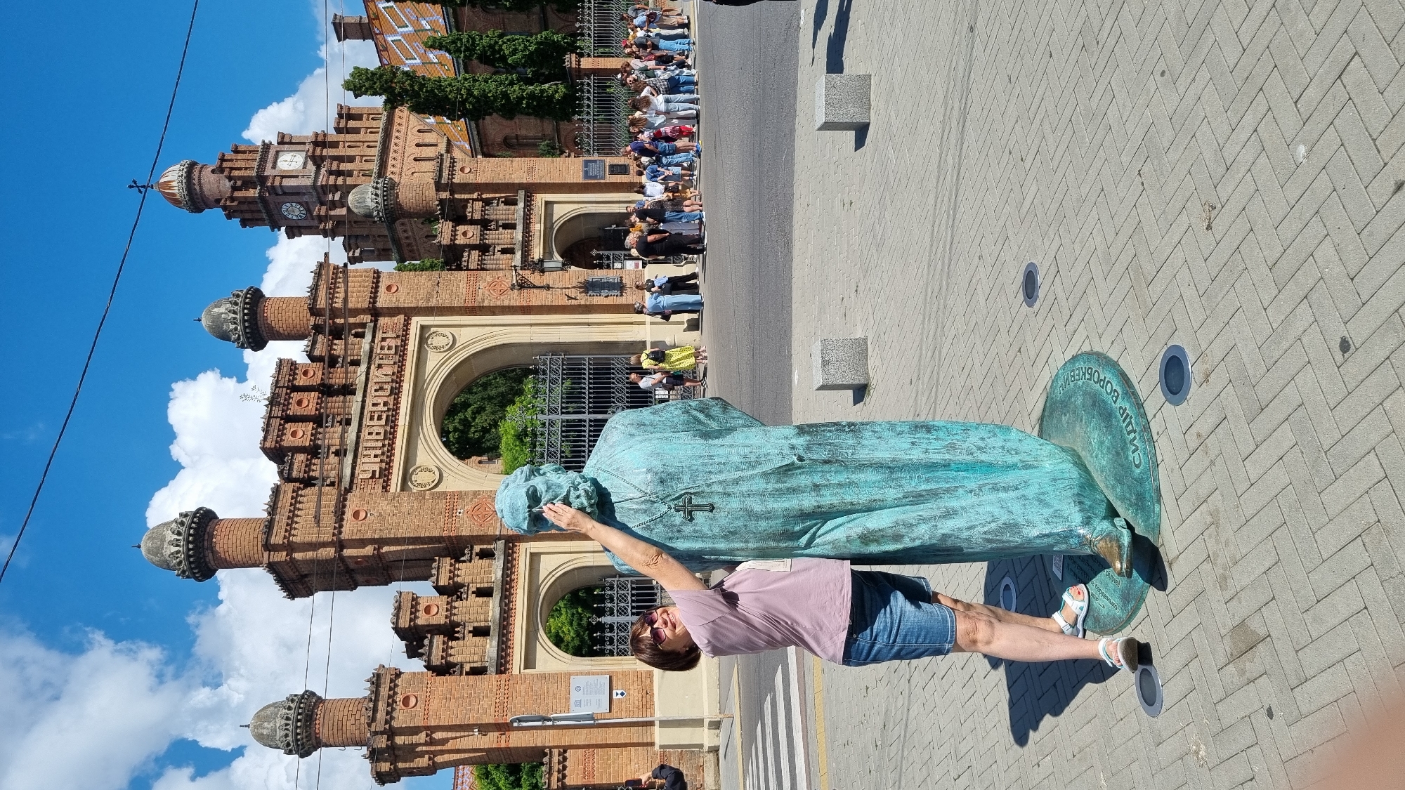 In Chernivtsi, Ukraine, a woman in a pink t-shirt and denim shorts stands in front of a statue of a man wearing a robe and a cross necklace. She is touching the statue's face with her right hand and smiling. The statue is a greenish-blue color and stands on a circular base with text. Behind them is the grand entrance of Chernivtsi National University, a UNESCO World Heritage site, constructed of red brick with ornate towers and an archway. A group of people are visible in the background, lined up near the entrance. The sky is blue with scattered clouds, and the ground is paved with bricks.