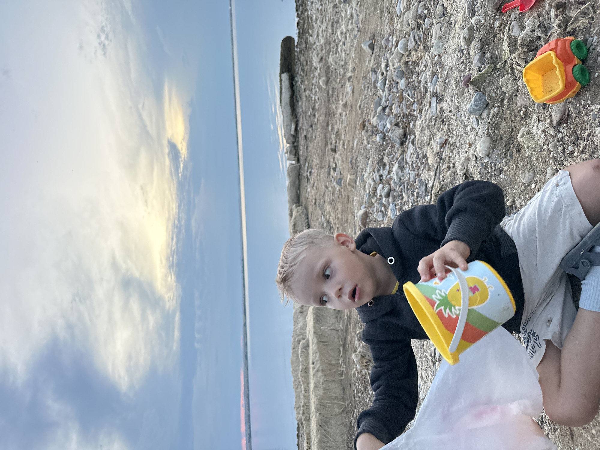 In Energetik, Russia, a young boy with blonde hair sits on a rocky shore by a body of water. He is wearing a black hoodie, light-colored shorts, and socks. In his left hand, he holds a white plastic bag, and in his right hand, he clutches a colorful yellow bucket decorated with a pineapple. Beside him, a small orange and green toy truck sits on the pebbles. The background features a wide expanse of water under a cloudy sky with hints of yellow and pink from what appears to be a sunset or sunrise. Along the water's edge, there are piles of debris or natural material.