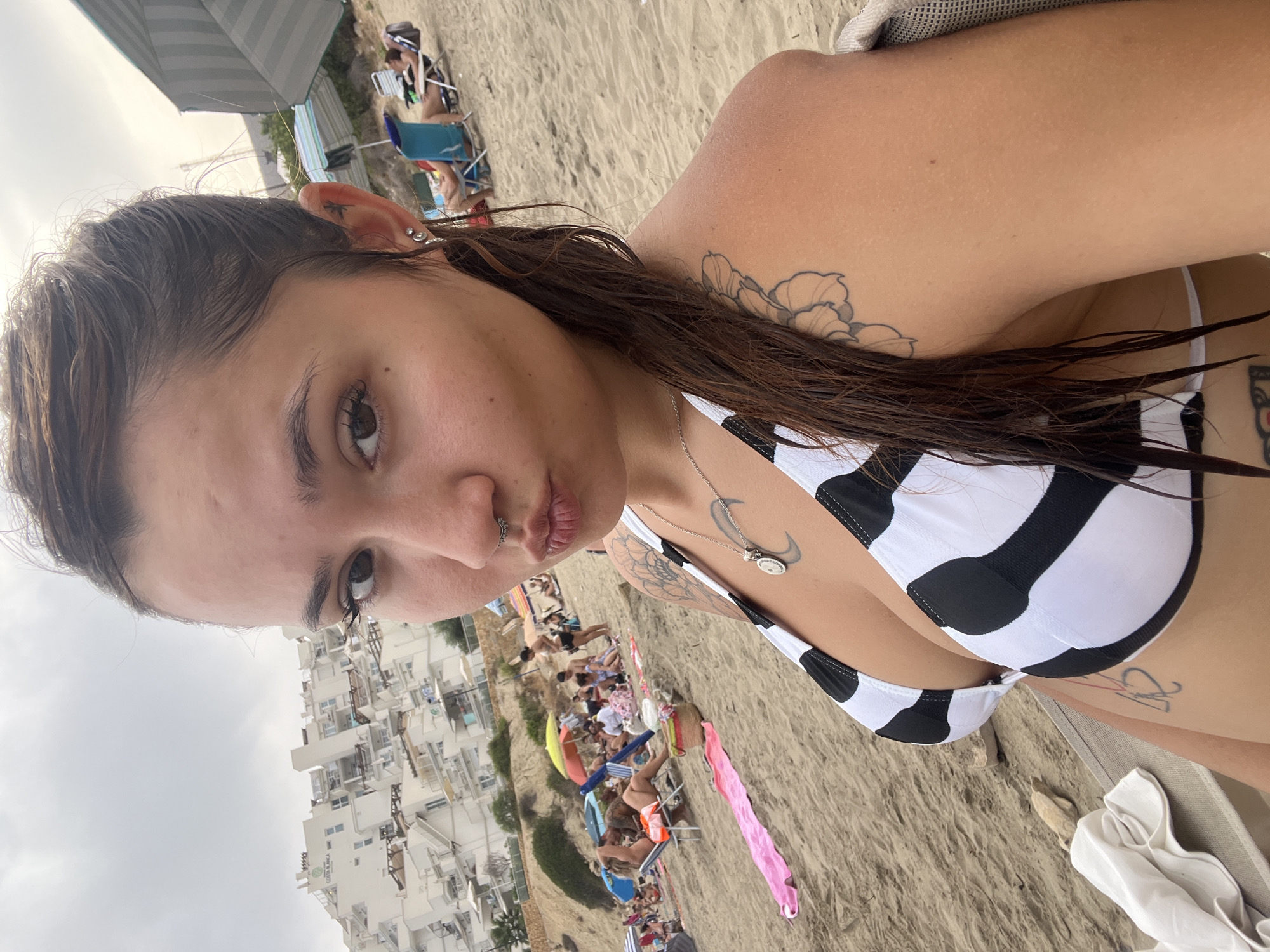 An outdoor, eye-level, medium selfie shows a young adult female with dark hair and tattoos on her shoulder and chest, wearing a black and white striped bikini top. She is on a sandy beach with other beachgoers and buildings in the background. The setting appears to be Urbanització Venta De Lanussa, Spain, based on the architecture of the buildings. The sky is overcast.