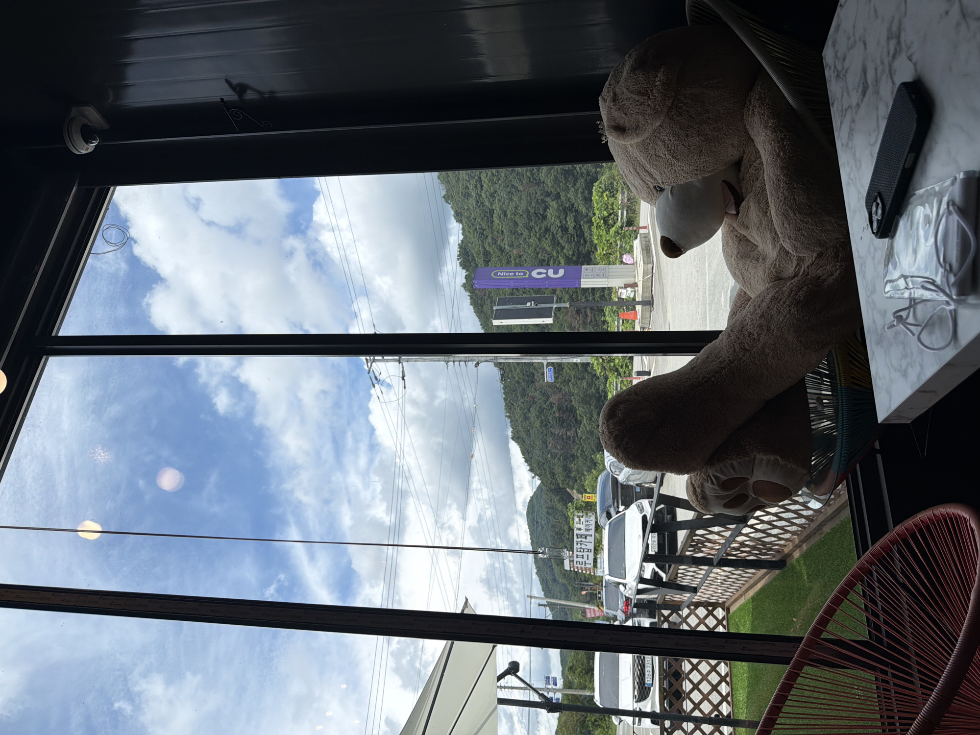 A large, plush teddy bear is seated in a chair indoors, facing a window. Through the window, a street scene is visible with parked cars and a green, hilly landscape under a cloudy sky. A purple sign with white text that reads "Nice to CU" is visible in the background, indicating the location is near a CU convenience store. On the table in front of the teddy bear are a cell phone and a clear plastic bag containing a white face mask.