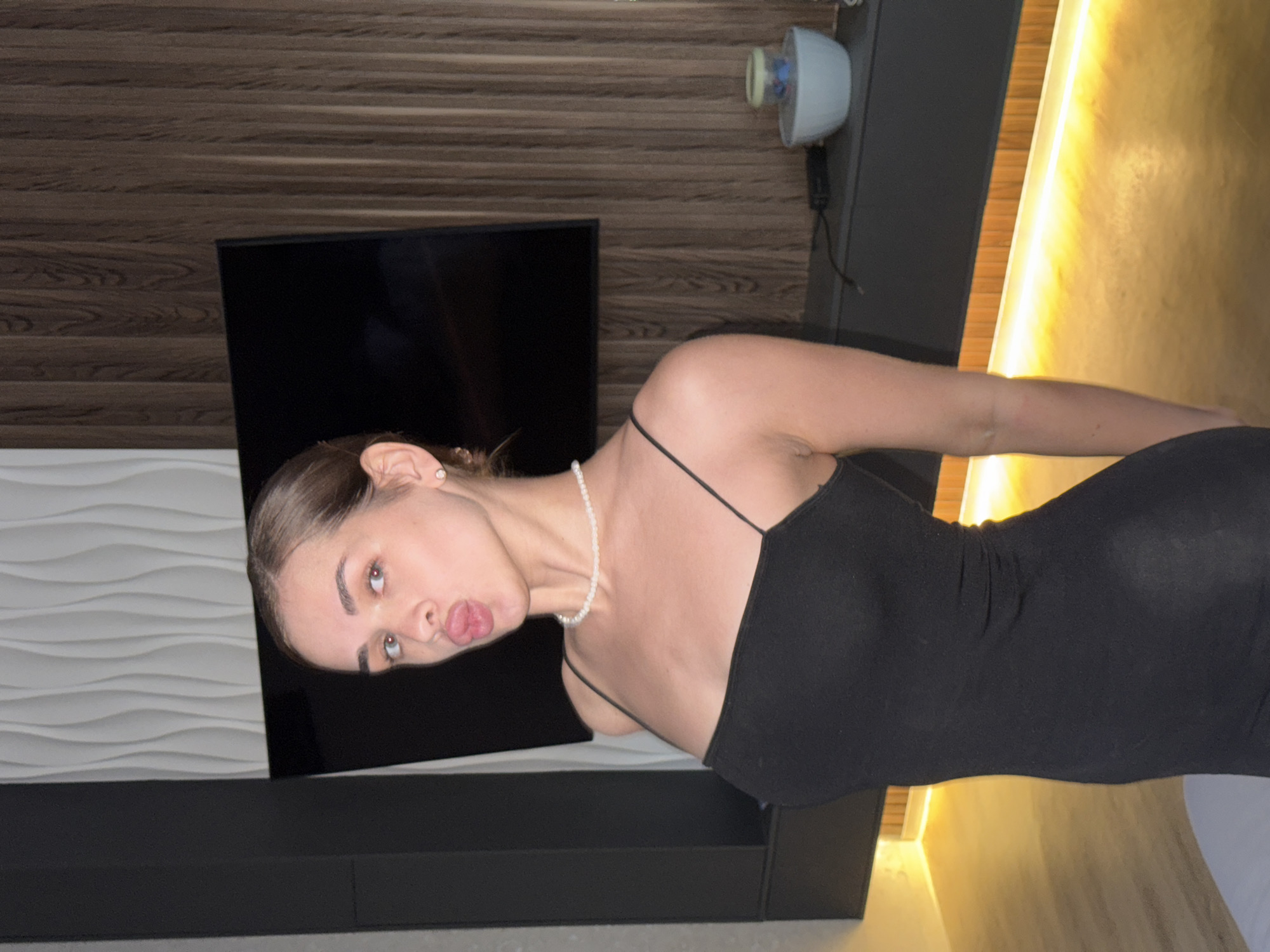 A woman in a black dress stands indoors. She is wearing a pearl necklace. Behind her is a wall with horizontal wooden slats and a black television. A white wavy panel is visible on the left. There is a cylindrical white object mounted on the wall. The location is indoors, but the city of Лобаново, Russia cannot be confirmed from the image.