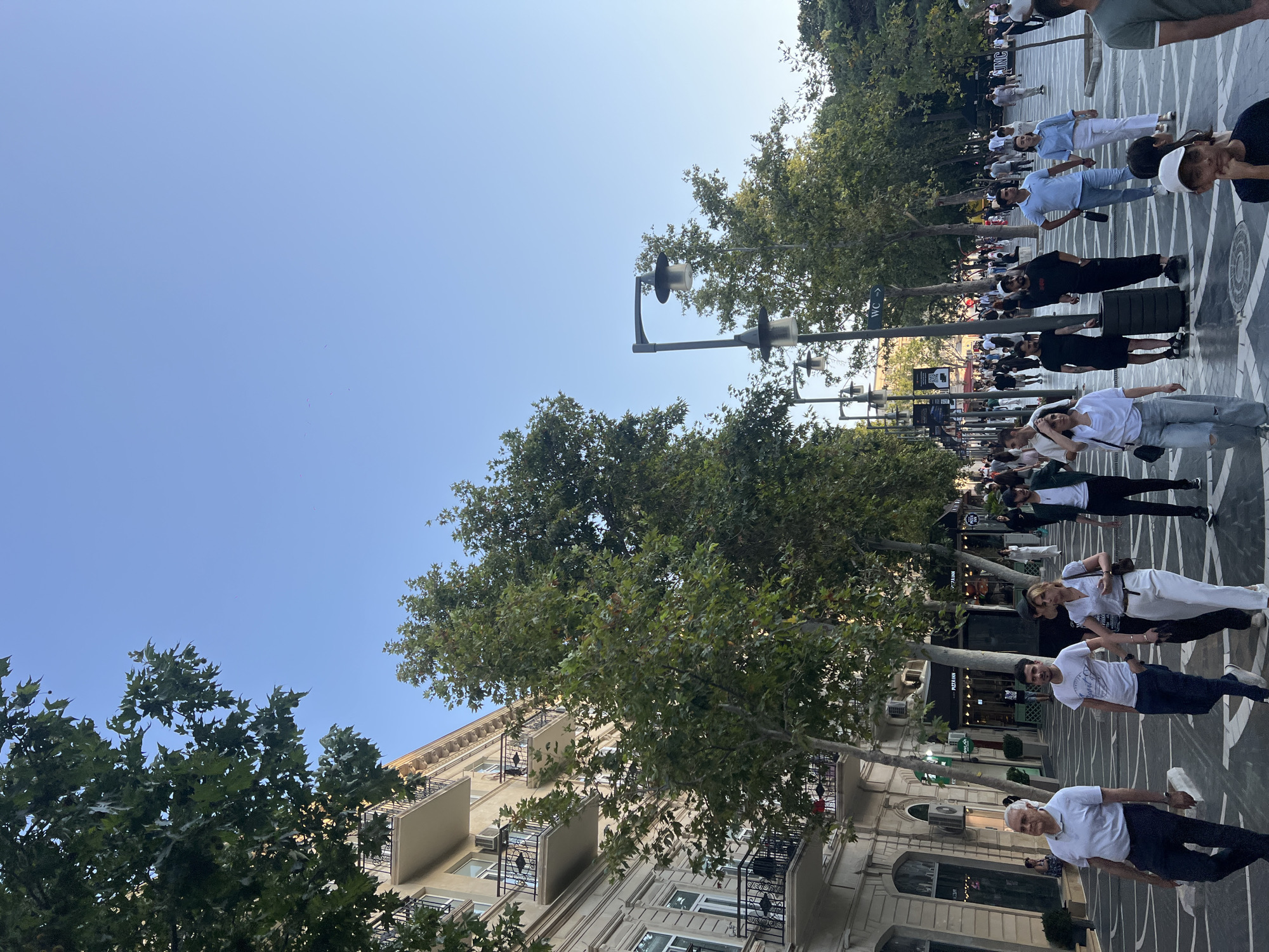 This image depicts a lively pedestrian street in Sabail Raion, Azerbaijan, likely Nizami Street, known for its shops and cafes. The scene is bathed in the warm light of late afternoon or early evening, casting long shadows across the patterned pavement. A diverse crowd of people is seen walking, talking, and browsing, creating a dynamic and engaging atmosphere.
In the foreground, a man in a white polo shirt and dark pants walks towards the left, while a couple in white clothing, a woman in a white blouse and white pants and a man in a light blue polo shirt, walks hand-in-hand towards the right. Behind them, a group of people are strolling along the street, with some pausing to look at storefronts. The architecture features elegant buildings with balconies and ornate detailing, interspersed with mature trees that provide shade and a natural element to the urban landscape. Streetlights, characteristic of the area, are visible along the walkway. Various shop signs, including "Pizza Inn," suggest commercial activity and dining options. The overall impression is one of a bustling city center, enjoying a pleasant day with many people out and about.
