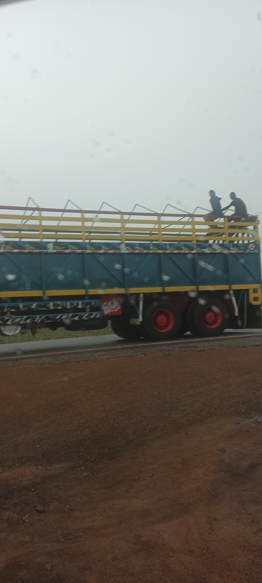 The image shows a blue truck with a yellow and red railing moving along a road. There are two individuals sitting on top of the truck. The tires of the truck are red. The road is bordered by brown dirt. The image quality is poor due to what appears to be raindrops on the camera lens. The location is Lafia, Nigeria.