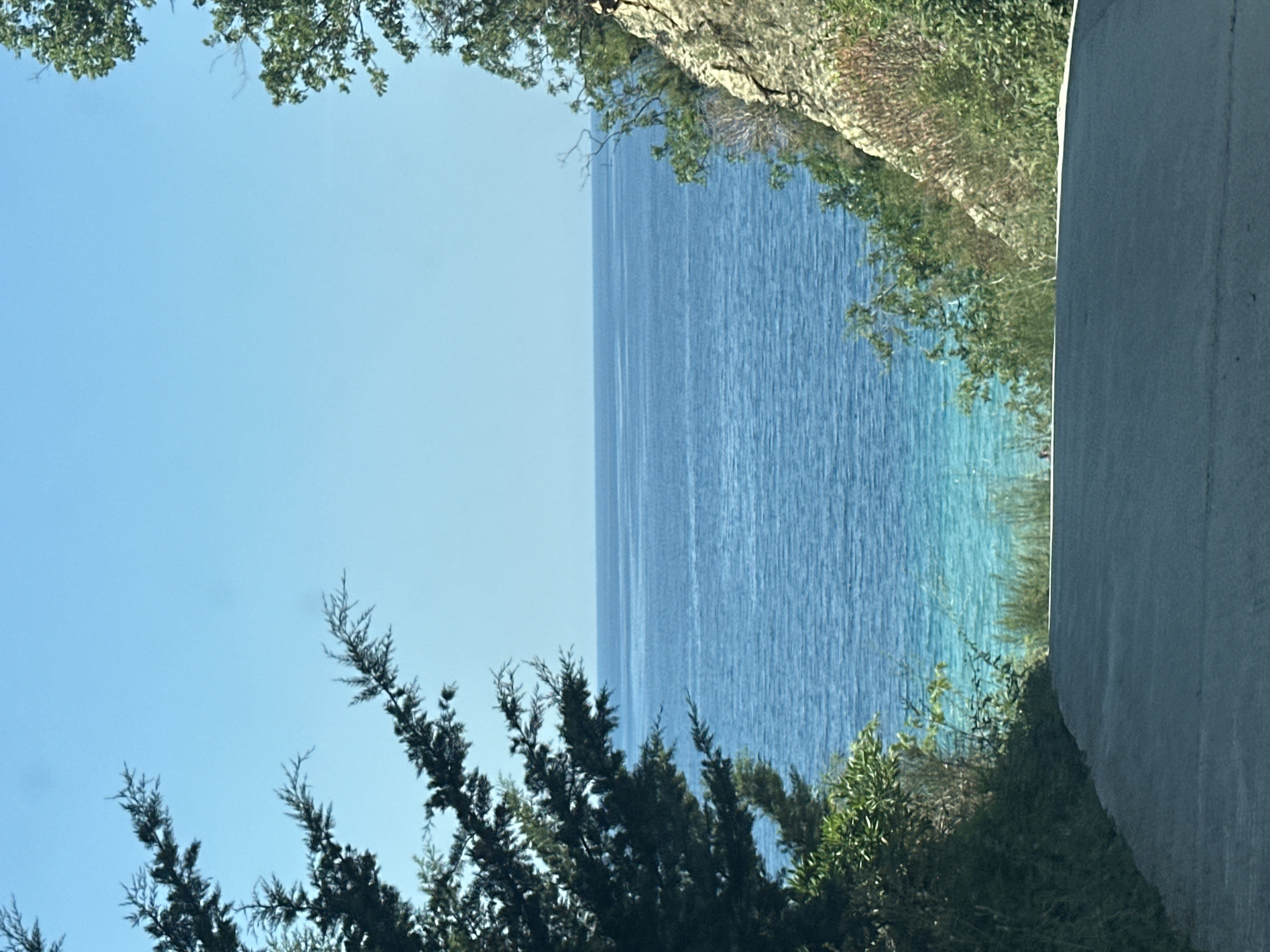 The image captures a serene natural scene, likely a coastal area. The main subject is a vast expanse of calm, blue water, reflecting the clear sky. The water transitions from a deeper blue further out to a lighter, turquoise hue closer to the shore, suggesting shallow depths or the presence of sand or vegetation beneath the surface.
Framing the water are elements of lush greenery and rocky terrain. On the top and left side of the image, leafy branches from trees with light green foliage overhang, creating a natural canopy. The right side of the image is dominated by a textured, light gray surface, possibly a wall or a paved area, which sharply contrasts with the organic elements. Below this, more dense green vegetation can be seen, leading down towards the water's edge.
The lighting suggests it is daytime, with a bright, clear sky. There are no people or specific objects clearly visible. The overall mood is peaceful and tranquil, emphasizing the beauty of the natural landscape. The image appears to be taken from a slightly elevated position, looking out over the water. The composition, with the natural elements framing the water and the stark contrast of the man-made surface, gives a sense of a secluded or protected viewpoint. There is no visible text in the image.
