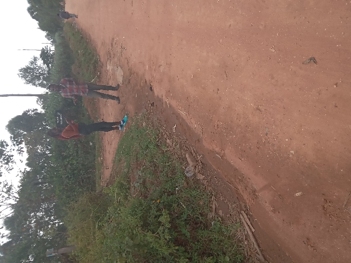 The image shows two people standing on a dirt road, located in Burundu, Kenya, at the coordinates (latitude: 0.491, longitude: 34.79). The woman on the left is wearing a long-sleeved orange shirt and dark blue jeans. She is looking at a sign that reads "Village Settlement" in black lettering. The man on the right is wearing a red and black plaid shirt and dark blue pants. He is looking at the woman. The background shows a dirt road, green vegetation, and trees. The scene appears to be a casual interaction between two individuals in a rural area.