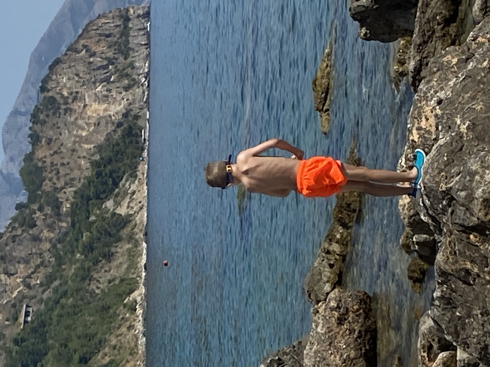 A young boy, seen from behind, stands on a rocky shoreline overlooking the sea. He is wearing orange swim trunks and blue swim shoes. A pair of goggles is pushed up onto his forehead. The sea is a deep blue, and in the distance, a rocky cliff face rises, covered in green vegetation. A small building is visible nestled on the side of the cliff. The sky above is clear and blue. The scene appears to be a natural coastal environment, likely in Prijevor, Montenegro, given the mountainous backdrop and the clear blue sea.