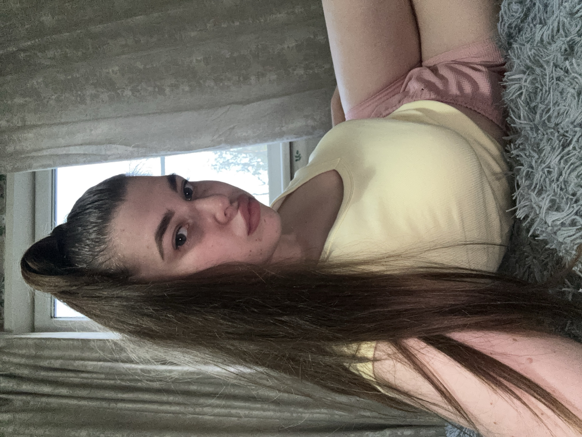 A woman with long, dark hair is lying on a gray, shaggy surface, possibly a bed or a blanket. She is wearing a yellow t-shirt and pink shorts. A window is visible in the background, suggesting an indoor setting. Curtains are partially obscuring the window. There are no indications of the location being Мещёры, Russia or any other city.