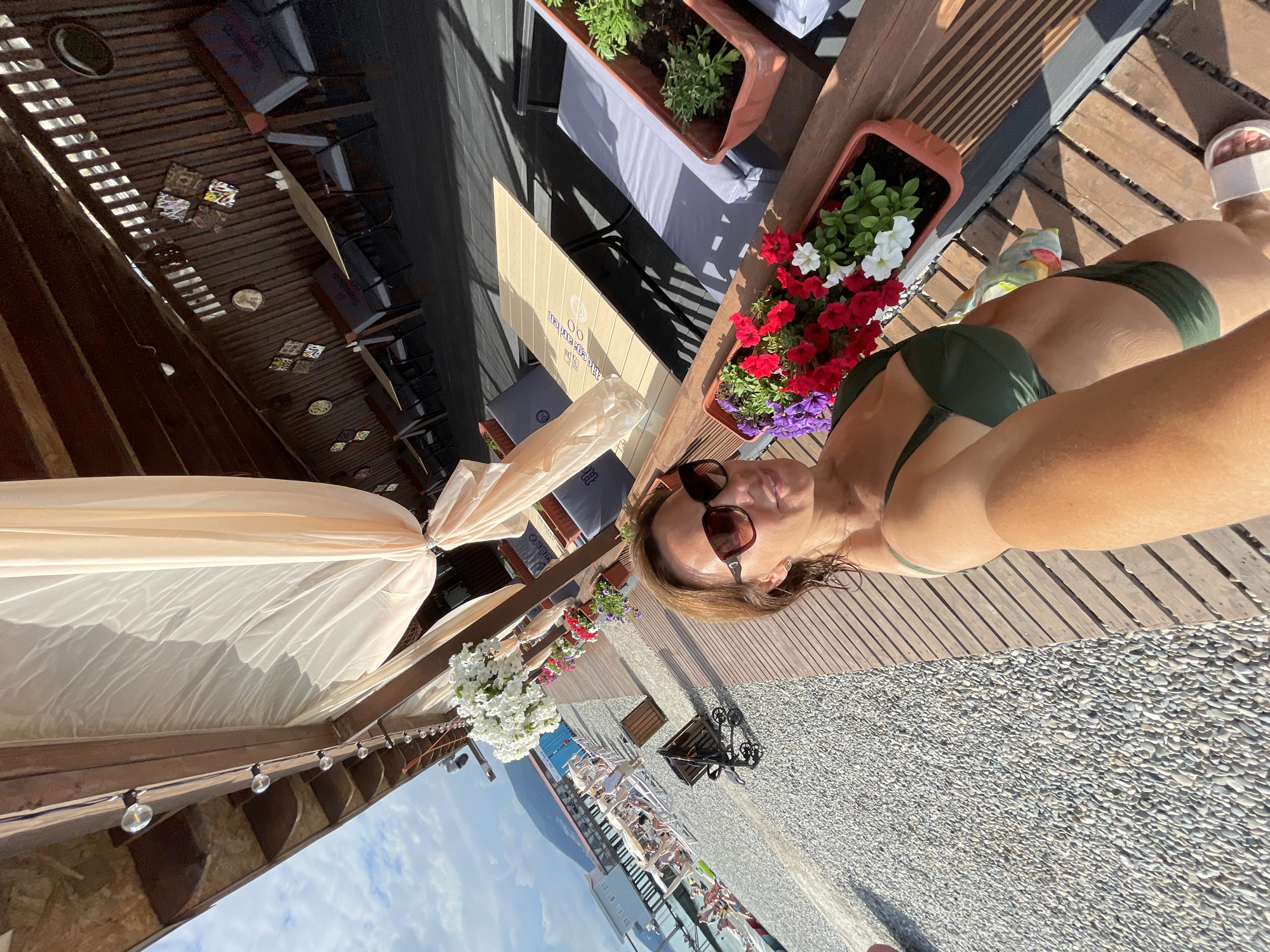 In a selfie taken from a high angle on a sunny day, a woman with shoulder-length brown hair and sunglasses is positioned on a wooden deck overlooking a pebble beach. She is wearing a dark green bikini. Behind her, a beach area with lounge chairs and umbrellas is visible, with people scattered throughout. Further in the background, a body of water and mountains can be seen, suggesting a coastal location. The deck features a wooden railing adorned with several planters filled with colorful flowers. A canopy overhead casts shadows on the scene. The overall impression is a resort or cafe setting in Sochi, Russia, likely near the Black Sea.