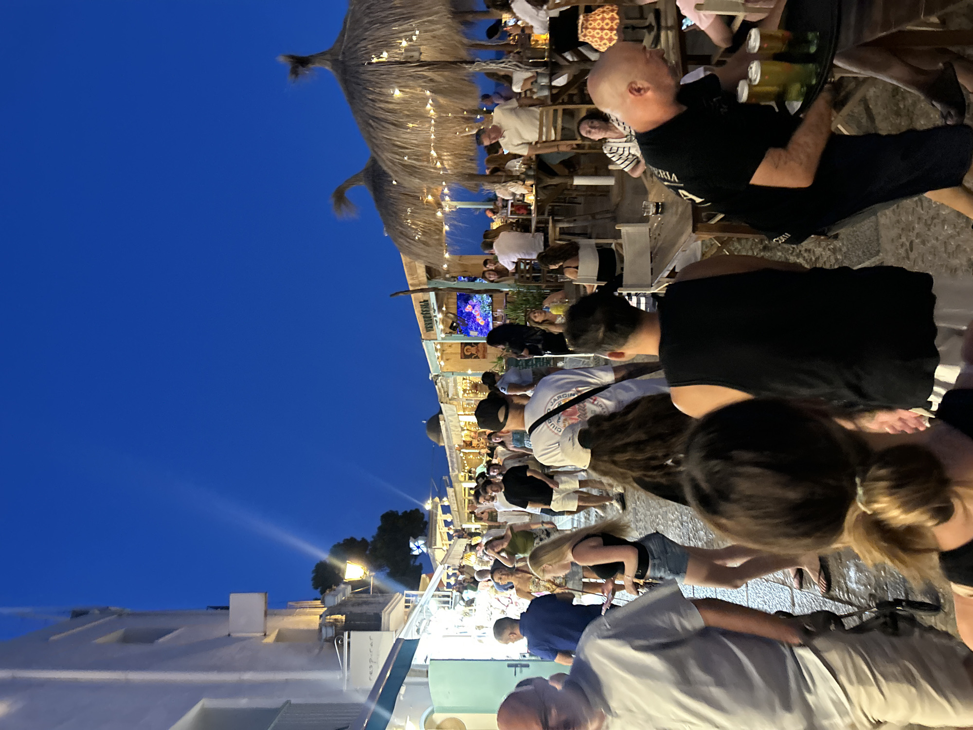 The image depicts a busy pedestrian street scene in Peníscola, Spain, at dusk. The sky is a deep blue, transitioning from day to night. On the left, a white building with lit windows and awnings lines the street. People are walking in both directions, many dressed in casual summer attire. Some are looking at their phones, while others are engaged in conversation.
To the right, several outdoor seating areas for restaurants and bars are set up, covered by thatched umbrellas. String lights are draped across the umbrellas, adding to the ambiance. People are seated at wooden tables, some eating and drinking. A waiter is visible carrying a tray with three glasses of beer. The ground is paved with cobblestones, which appear wet, suggesting recent rain or street cleaning. The overall atmosphere is lively and social, characteristic of a tourist area during the evening.