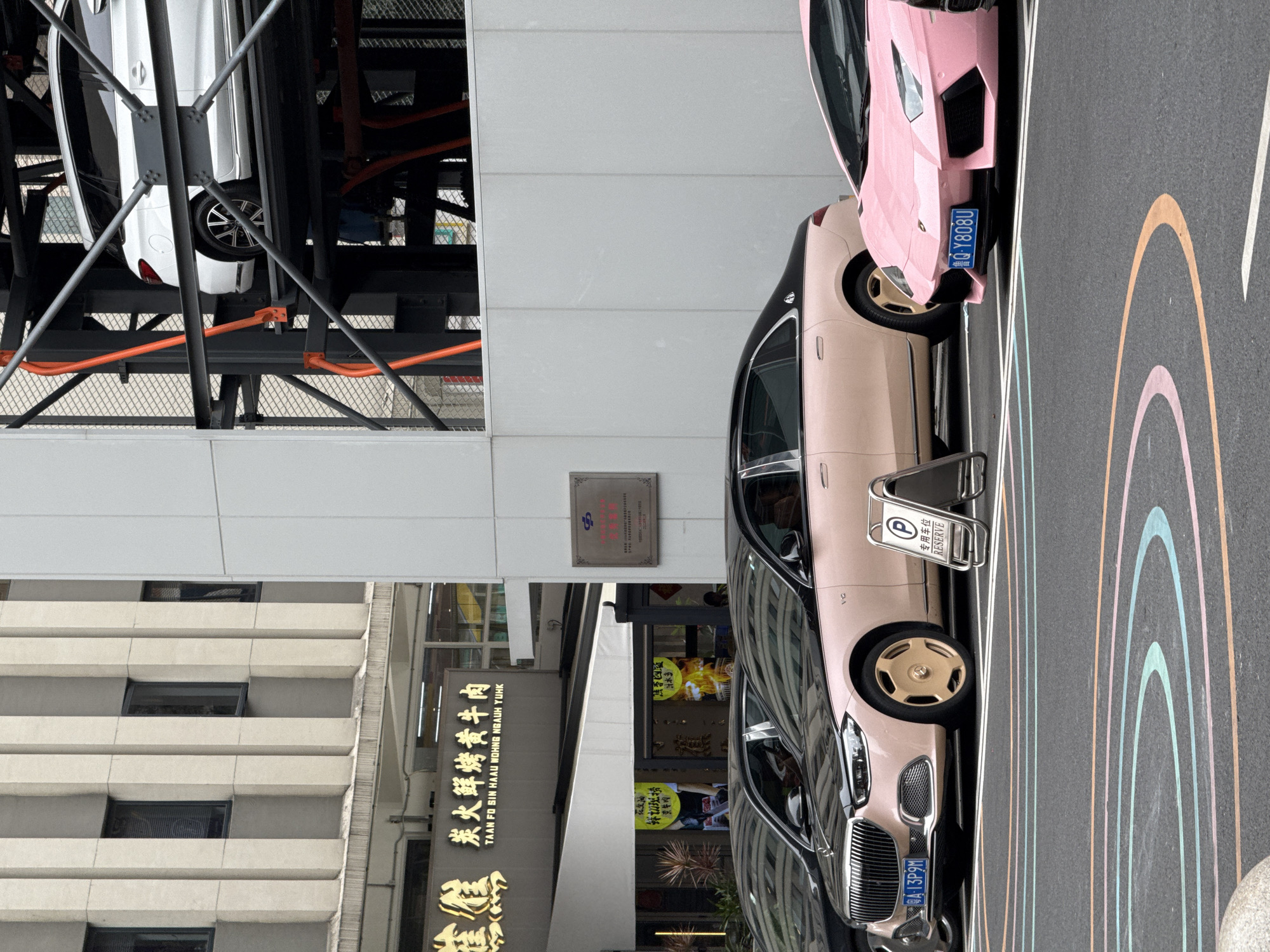 The image depicts a street scene in the Liwan District of China. A white multi-story building stands to the left, while a mechanized parking structure with a white car inside is visible. A sign for "Taan Fo Sin Haau Wohng Ngauh Yuhk", a restaurant specializing in charcoal grilled beef, is prominent. Two luxury cars, a champagne-colored sedan and a pink Lamborghini, are parked next to each other, with parking signs indicating reserved spots. A painted rainbow pattern adorns the parking surface. Chinese license plates are visible on the cars.