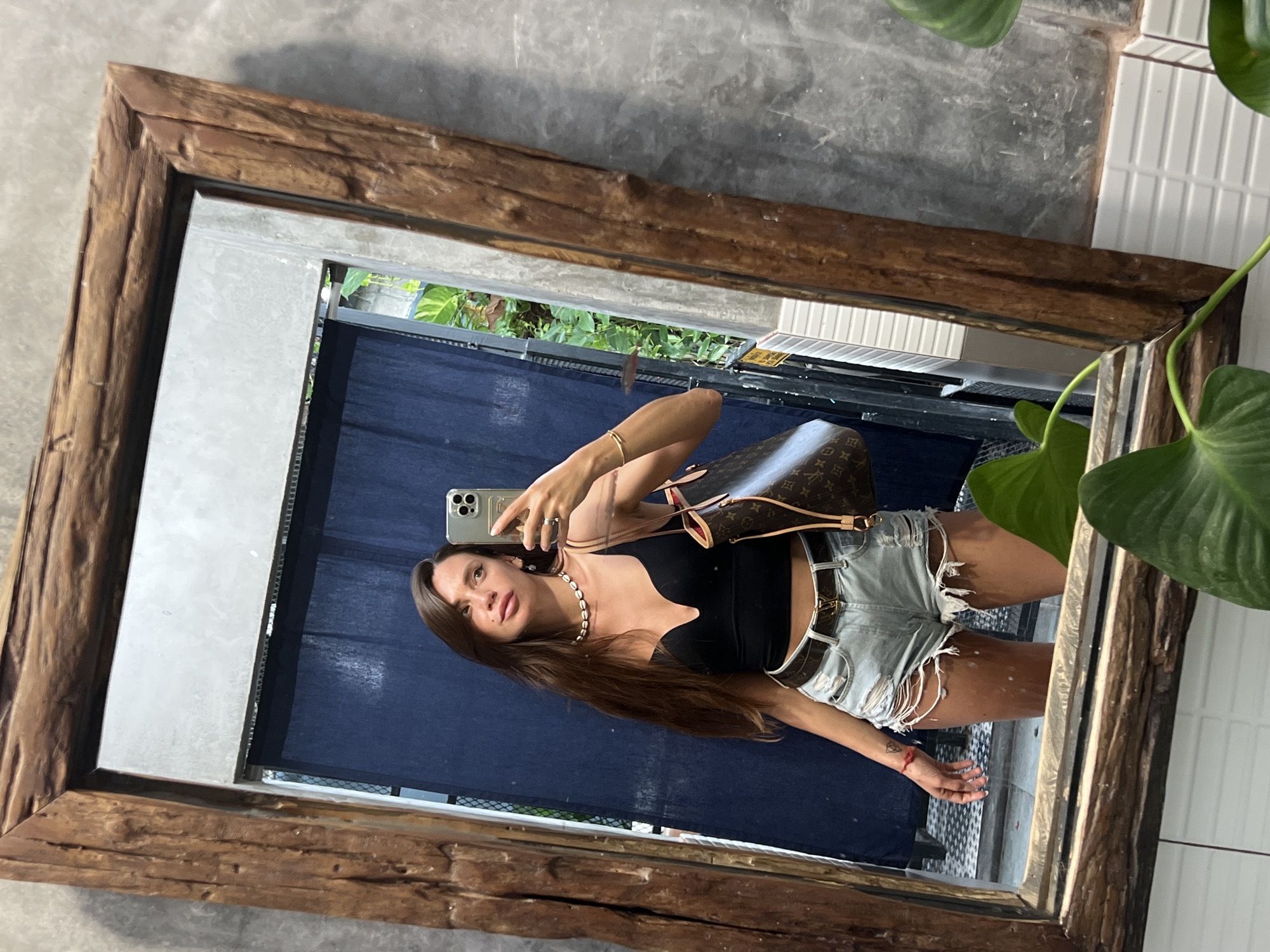 A full-shot, medium-angle, mirror selfie shows a woman standing in front of a blue curtain and a concrete wall. She has long brown hair and is wearing a black crop top and distressed denim shorts. She is also wearing a belt with a gold buckle, a shell necklace, and a gold bracelet. In her right hand, she is holding a smartphone and a designer handbag with a brown and tan monogram print is slung over her left shoulder. The mirror is framed with rustic, dark wood. Green plants are visible in the foreground and background.