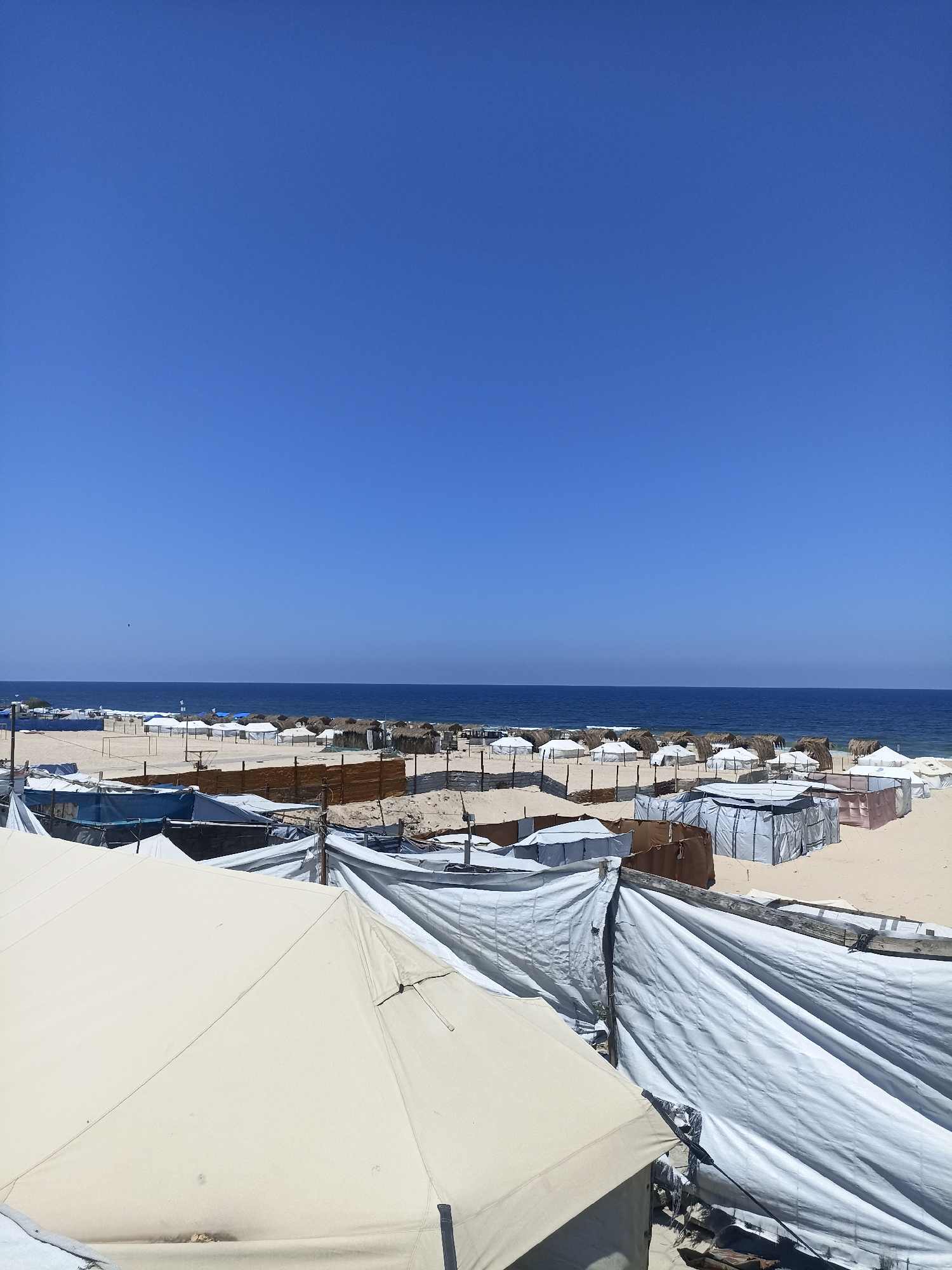 The image depicts a beach scene in Gaza under a clear blue sky, suggesting a warm, sunny day. In the foreground, various tents and temporary shelters made of white and beige tarpaulin and some with straw roofs are visible, arranged on the sandy beach. These structures appear makeshift and are densely packed. Further in the background, the calm blue ocean meets the horizon. Beyond the beach, more structures and what looks like a fence are present. There are no people visible in the image. The overall impression is one of a temporary encampment or settlement on a beach. The time of day is likely midday due to the bright lighting and clear sky. The location is identified as Gaza, but the specific site is unknown. There is no visible text in the image.