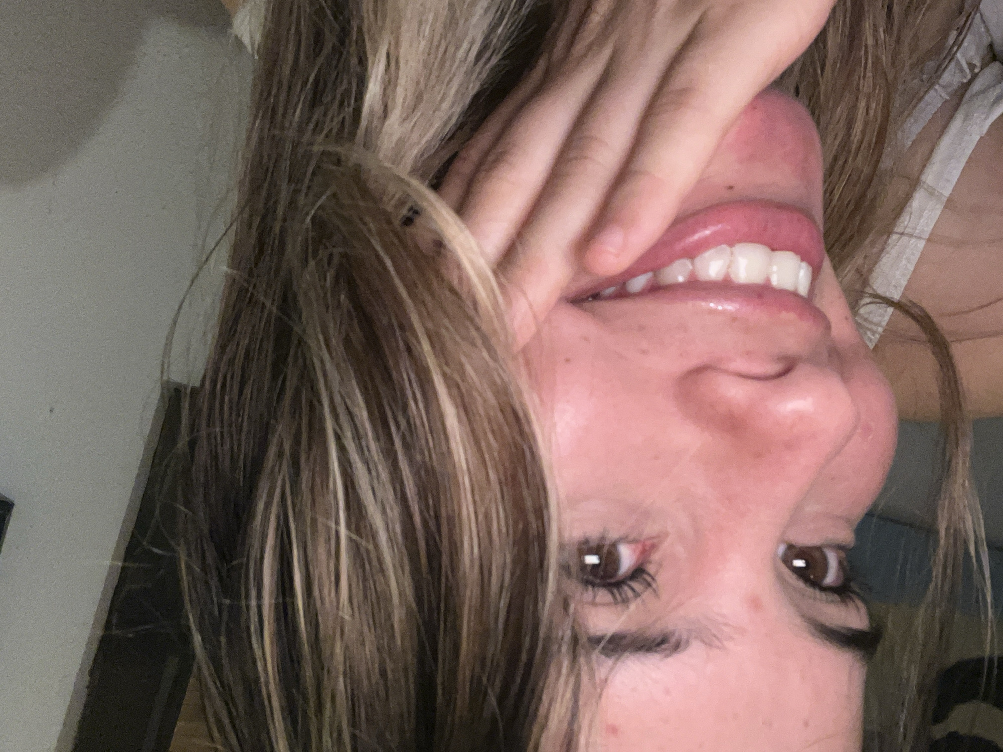 A close-up, upside-down selfie of a fair-skinned person with brown eyes and light brown hair with blonde highlights. The person's right hand is covering part of their face, near the mouth. They are smiling, revealing white teeth and pink lips. A small portion of a white tank top is visible on the right side of the frame. The background is blurry, suggesting an indoor setting, with glimpses of a white wall and a dark object. It is impossible to determine the location of Sumalao, Argentina from this image alone.