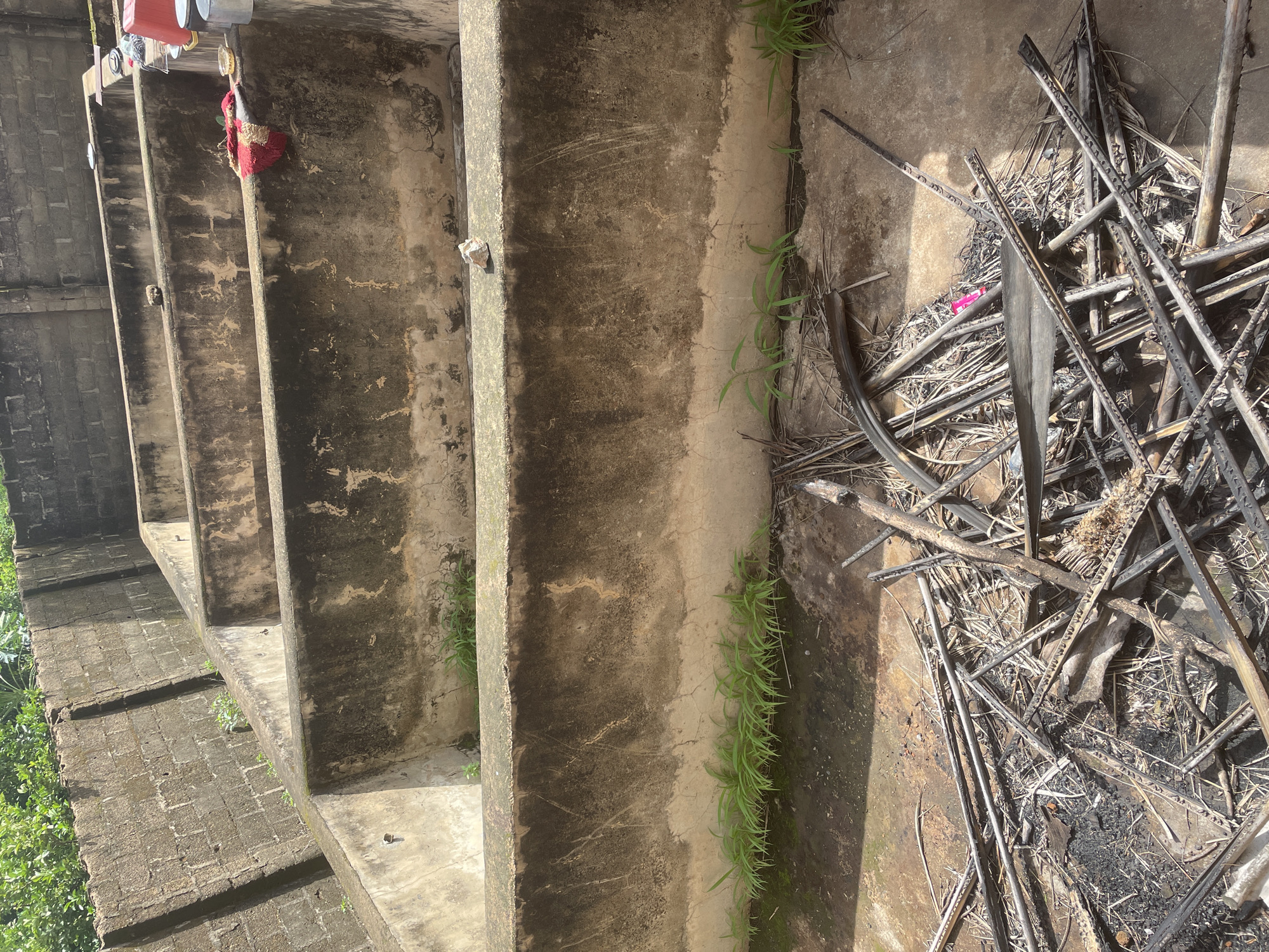 In Kakau, Nigeria, a series of tiered concrete basins are visible, possibly remnants of a former industrial or agricultural facility. The basins are weathered and show signs of disrepair, with some vegetation growing along their edges. In the foreground, a pile of dry, burnt vegetation and debris lies on the concrete floor, suggesting recent or past fires. A red cloth is draped over the edge of one of the basins in the middle ground, and various containers, including a red plastic basin and metal bowls, are placed on the edge of the furthest basin. The background features a brick wall and lush green foliage, indicating an outdoor setting with some overgrowth. The scene appears to be an abandoned or neglected area within the city.