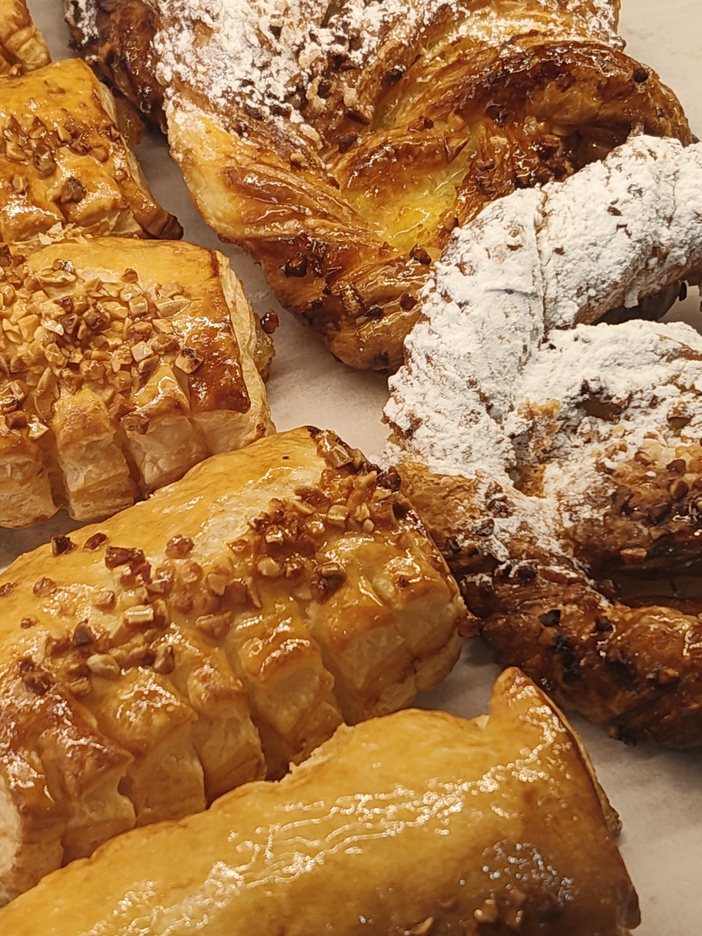A high-angle close-up photograph reveals an assortment of pastries on a white surface. The pastries are various shapes and sizes. Some are square or rectangular with layered edges, covered in chopped nuts and a glossy glaze. Others have twisted, braided shapes with visible layers, some dusted with powdered sugar. The lighting is bright and even, highlighting the textures and colors of the baked goods. The location is likely a bakery or pastry shop in Dénia, Spain. The image focuses on the pastries; there are no discernible people or specific interactions. The environment consists of the pastries on the white surface.