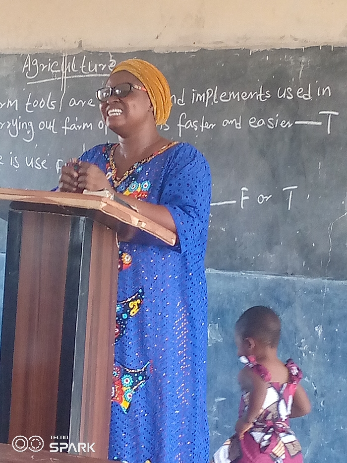 The image depicts an indoor scene in Ajaokuta, Nigeria, likely a classroom or lecture setting. In the foreground, an adult woman is positioned behind a wooden podium. She wears a yellow headwrap, black-rimmed glasses, and a blue long-sleeved dress adorned with colorful, intricate patterns. Her hands are clasped on the podium's upper surface, and she displays a broad smile while looking towards her upper right.
Behind the woman, a dark chalkboard is visible, featuring white chalk writing. The prominent heading reads "Agriculture." Below this, text states: "farm tools / are used and implements used in carrying out farm operations faster and easier — T". An incomplete line further down reads "is use f ______ F or T ." The wall above the chalkboard is a light, neutral color.
A young child, appearing to be a toddler, is partially visible in the lower right portion of the frame. The child has short, dark hair, is wearing a multi-colored patterned dress predominantly featuring red/maroon hues, and is facing away from the camera, seemingly in motion. A "TECNO SPARK" watermark is present in the lower-left corner of the image.
