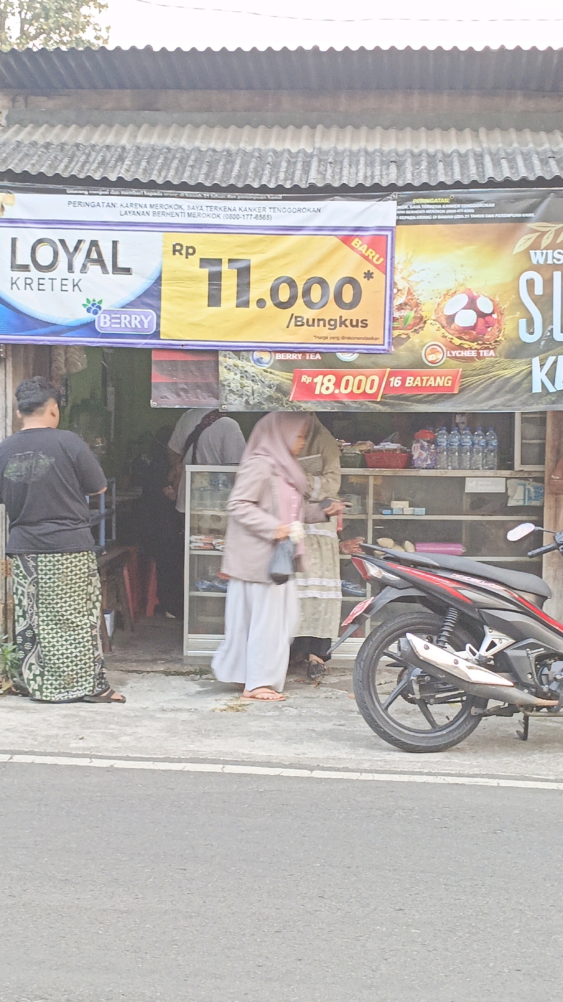 A street-level, medium-shot photograph captures a street-side stall in Salatiga, Indonesia. In the foreground, a grey asphalt road with a white line runs horizontally across the bottom of the frame. To the right, the rear section of a red and black motorcycle is parked. The stall itself is set up against a building with a corrugated metal roof and white-washed walls.
Several people are present. On the left, a man wearing a black t-shirt and a green and white patterned sarong stands with his back to the camera, facing the stall. In the center, a woman wearing a light pink hijab and a beige jacket over a long white skirt stands facing right, appearing to interact with the stall. Behind her, another person in a light-colored shirt and a dark head covering is visible inside the stall.
The stall features a glass display counter filled with various items, including bottles of water and what appear to be packaged goods. A red basket is visible on a shelf within the stall. Behind the counter, the interior of the building is dimly lit, revealing more shelves and products.
Above the stall, large banners are displayed. A white banner at the top contains Indonesian text and an image of a cigarette pack with a health warning. Below this, a bright yellow banner advertises "LOYAL KRETEK" with the price "Rp 11.000 / Bungkus" and a berry logo. To the right of this, another banner displays "WIS" and part of a word that is obscured, with an image of lychee tea. Further to the right, a red banner displays "Rp 18.000 / 16 BATANG" and mentions "BERRY TEA". The overall scene suggests a typical Indonesian street vendor selling cigarettes and other convenience items.