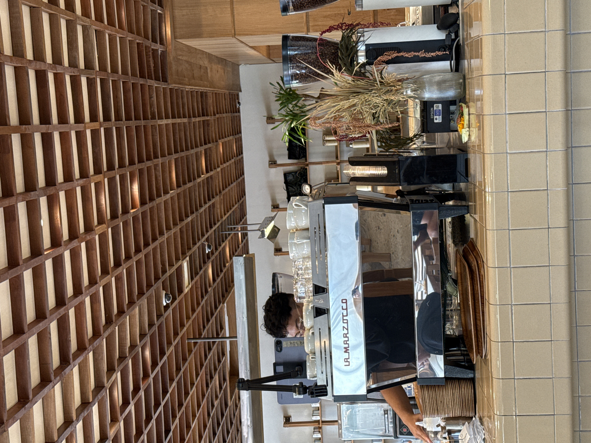 The image captures an interior scene, likely within a café or coffee shop located in North Kuta, Indonesia. The scene is dominated by a wooden grid-like structure covering a large portion of the wall on the left side of the frame. In the foreground, a person is seen operating a professional espresso machine, identifiable by the "La Marzocco" branding. The person appears to be in the process of preparing a beverage. The espresso machine is surrounded by cups, tools, and related accessories. Behind the espresso machine, decorative plants and coffee beans are displayed on shelves, adding to the aesthetic of the space. The right side of the image features a tiled wall.