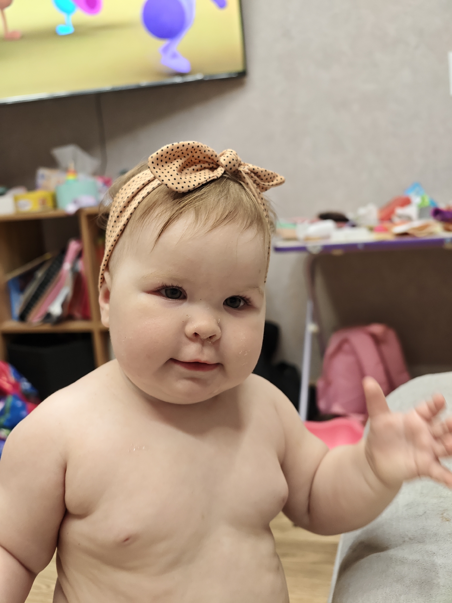 A baby with a polka dot headband is facing the camera with their right arm extended. The baby has fair skin and appears to be standing in a room with cluttered shelves and a television in the background. The location is Chaikovsky, Russia.