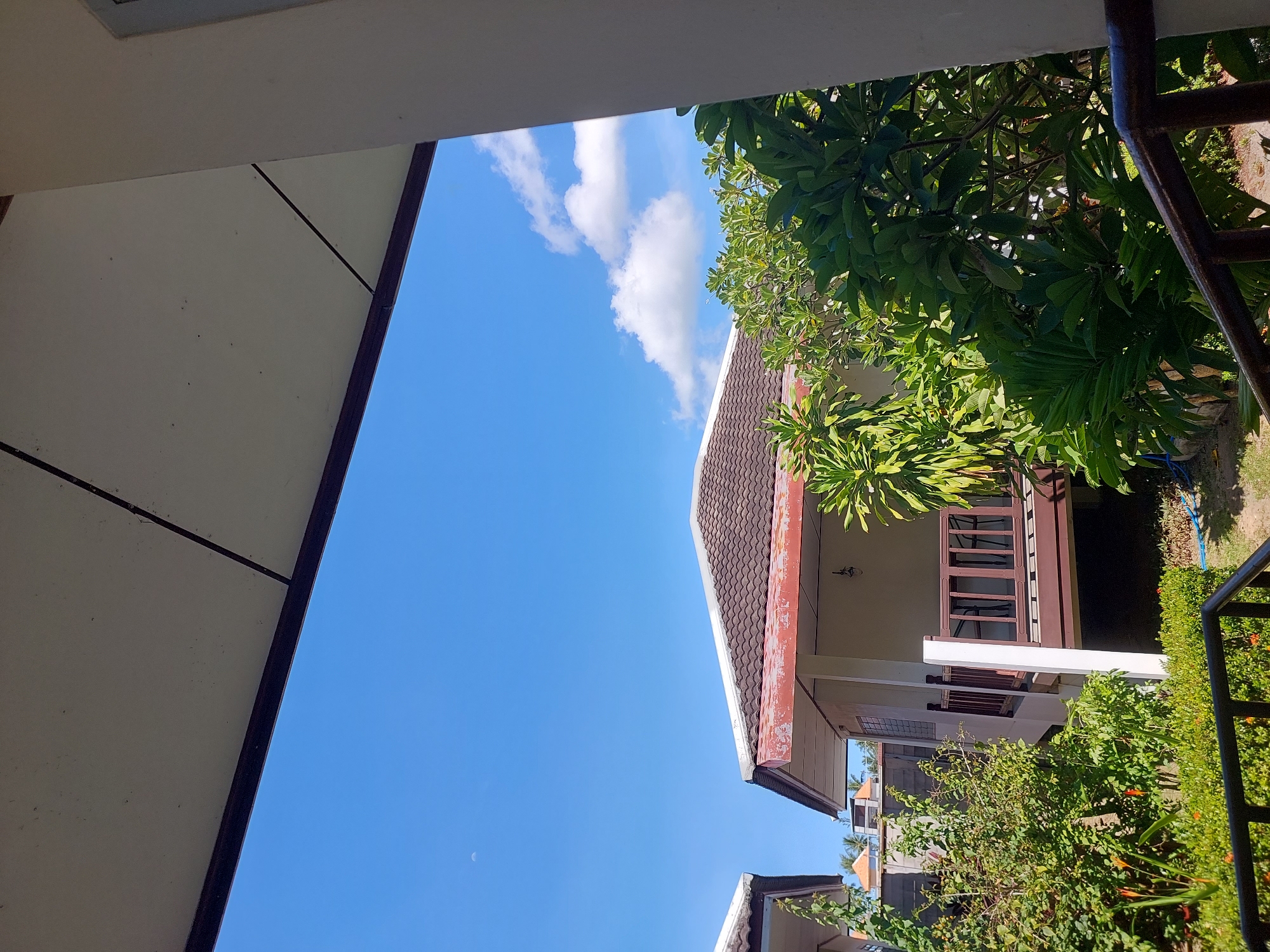 The image depicts a view of a tropical resort or residential area in Baan Mae Nam, Thailand. In the foreground, the edge of a white ceiling or overhang is visible, with dark beams outlining its structure against a clear blue sky. Below this, a small bungalow-style house is partially visible, featuring a brown tiled roof and a porch with a wooden railing. Lush green tropical foliage surrounds the house, with large-leafed plants and some flowering plants with orange blossoms. The sky is bright blue with a few scattered white clouds and a faint crescent moon. In the distance, the roofs of other similar buildings are visible, along with what appear to be palm trees, suggesting a coastal location. A dark metal railing is present in the lower part of the frame, indicating a balcony or staircase. The overall scene is bright and sunny, characteristic of a tropical climate.