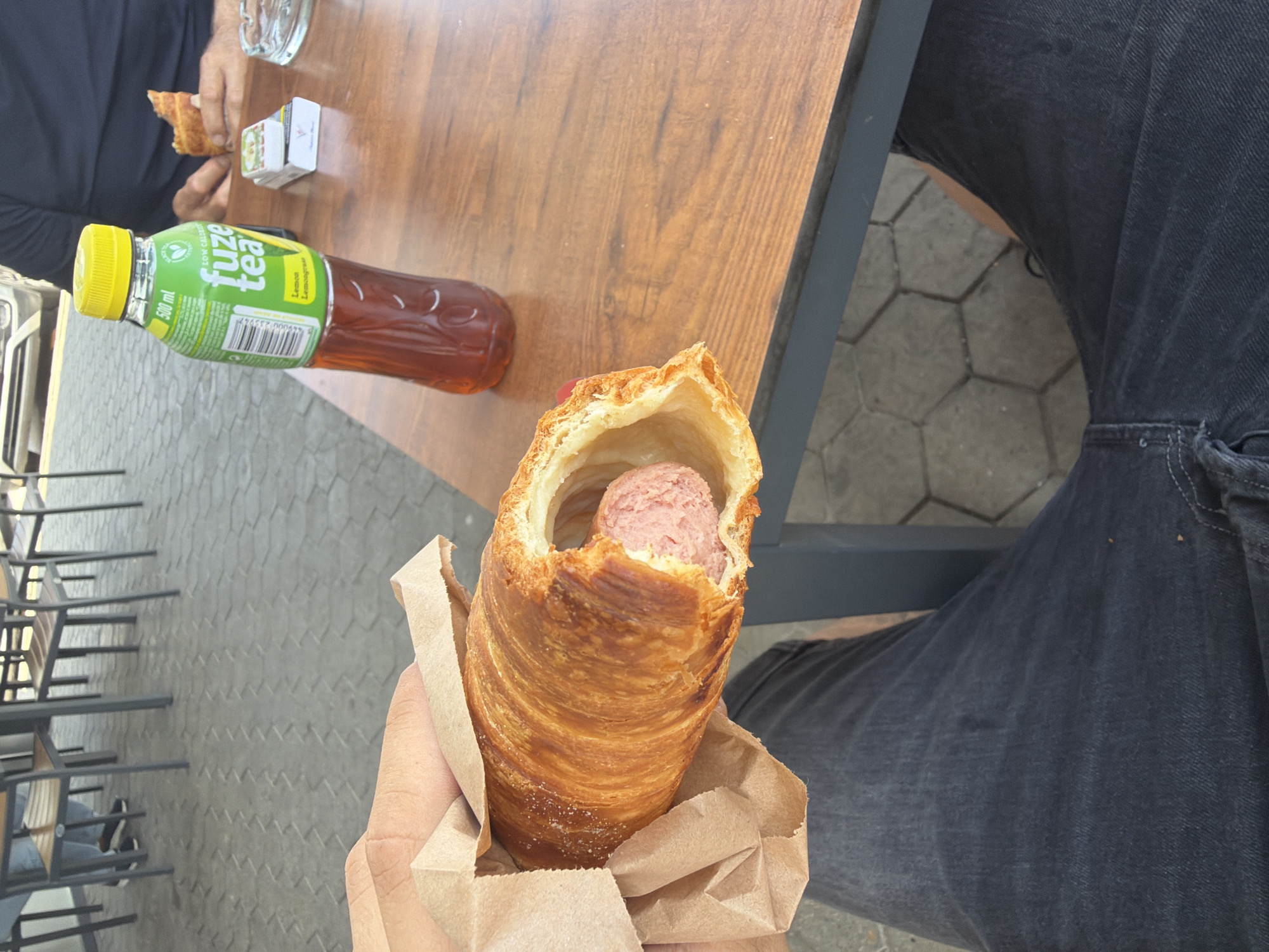 The image captures an outdoor dining or snack scene, likely at a casual eatery in Osikovitsa, Bulgaria. The main subject is a hand holding a large, golden-brown pastry, possibly a cronut or layered croissant, with a pink-colored sausage peeking out from its hollow center. The pastry is served in a brown paper wrap.
In the background, a wooden table holds a bottle of Fuze Tea with a bright green label and a yellow cap. The label clearly shows "Fuze Tea," "Lemon Lemongrass," "500 ml," and "LOW CALORIES," along with a barcode. Next to the bottle, another person's hands are visible, holding a piece of what appears to be a croissant or similar pastry. An ashtray and a small white pack of cigarettes are also on the table.
The foreground shows the leg of a person wearing dark-colored jeans. The ground beneath the table and chairs features grey hexagonal pavement tiles, indicating an outdoor setting. Dark metal frames of chairs are also visible. The bright, clear lighting suggests it is daytime, likely midday or afternoon. No specific emotions are discernible from the people in the image.