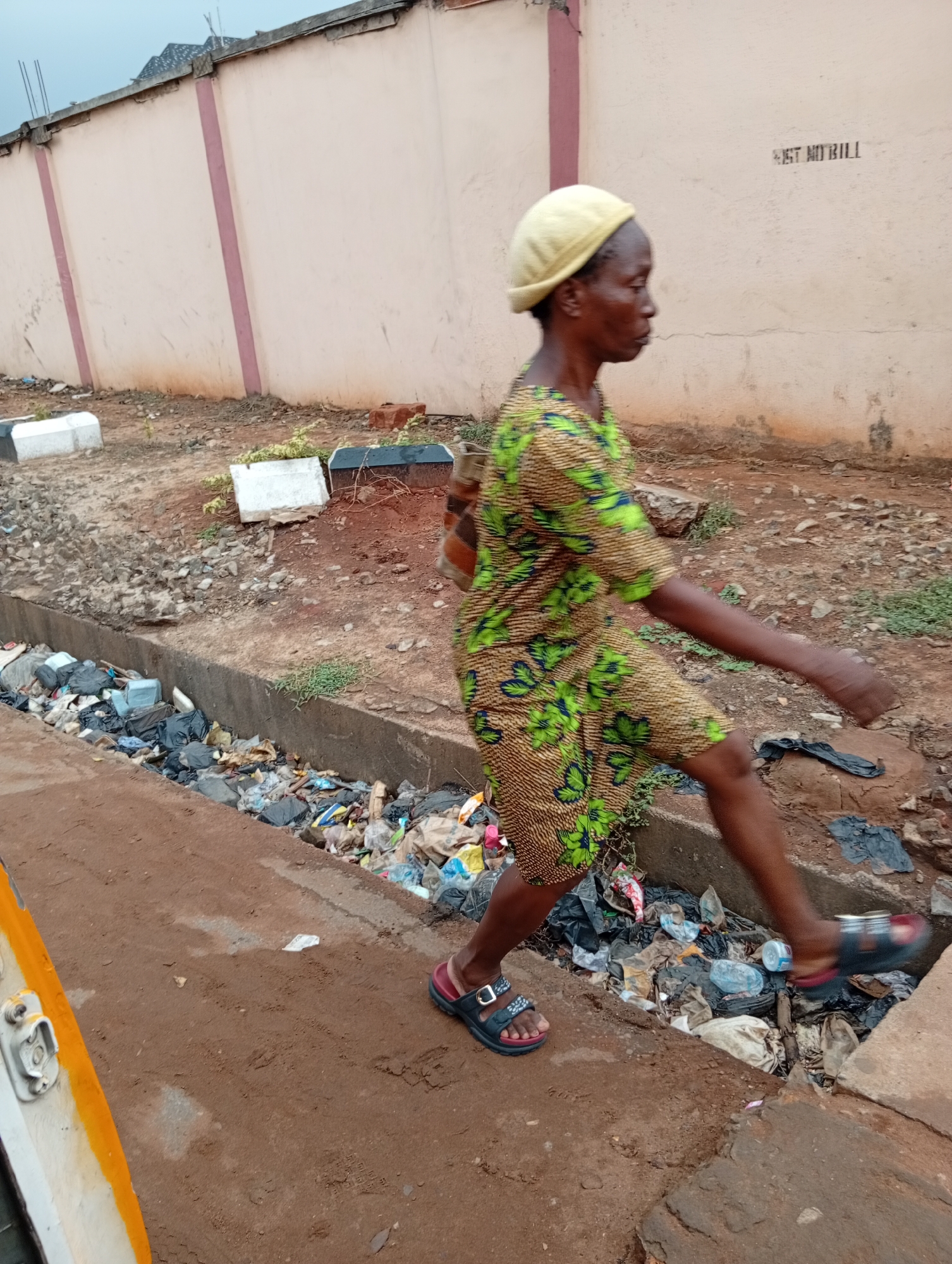 An older woman, wearing a yellow hat and a green and yellow patterned dress, is walking across a narrow, concrete-lined drainage ditch filled with trash in Ogidi, Nigeria. She is wearing sandals. The ditch is located next to a dirt path, and a painted wall runs in the background. The writing "WEST N'ODILI" is visible on the wall.