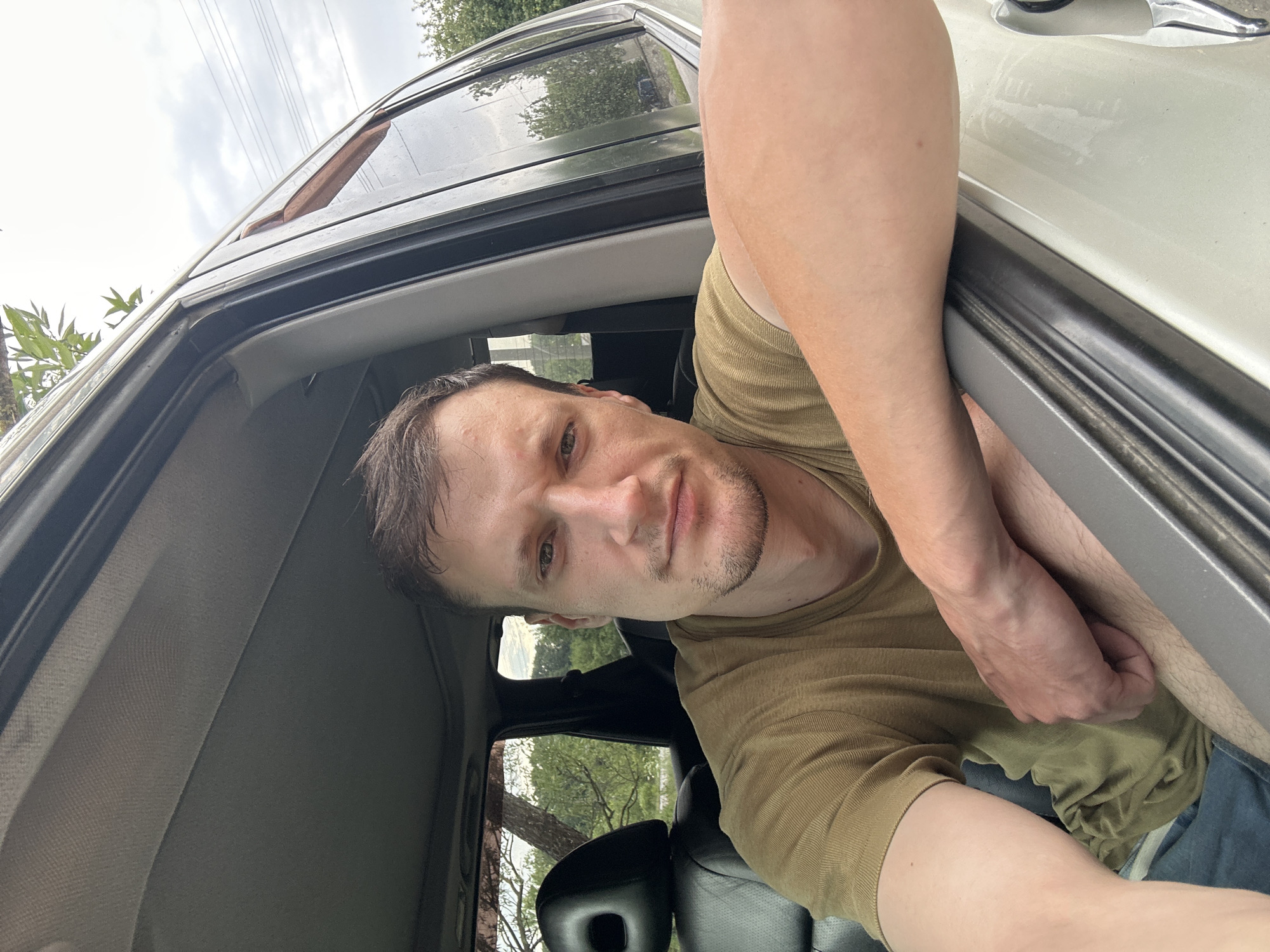In Makiv, Ukraine, a man with light brown hair and stubble is pictured from the chest up, seated inside a light-colored vehicle. He is looking directly at the camera, with his left arm resting on the open car door frame. He is wearing a light brown t-shirt and blue jeans. The interior of the car, including the black leather seat and the roof lining, is visible. Outside the car, trees and a cloudy sky can be seen.
