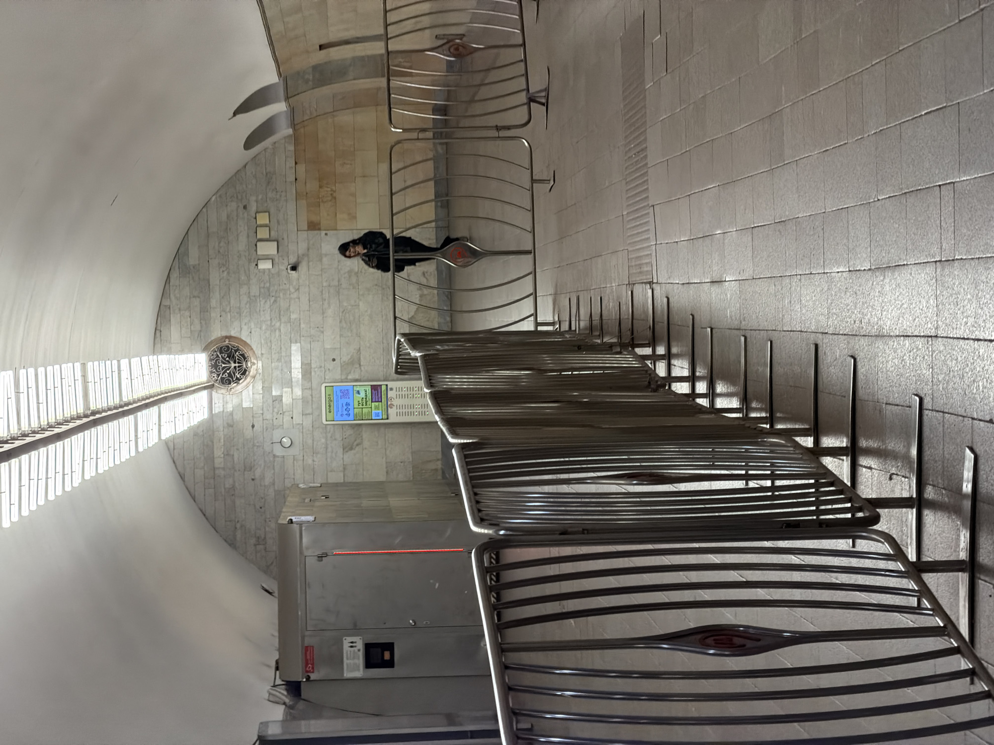 The image shows the interior of the Kiyevskaya metro station in Moscow, Russia. The architectural style is characterized by vaulted ceilings and tiled walls. A woman is standing near a row of metal barriers. An information screen is visible on the wall near a ticket vending machine. Sunlight is streaming in through windows along the ceiling. The overall environment appears to be clean and well-maintained.