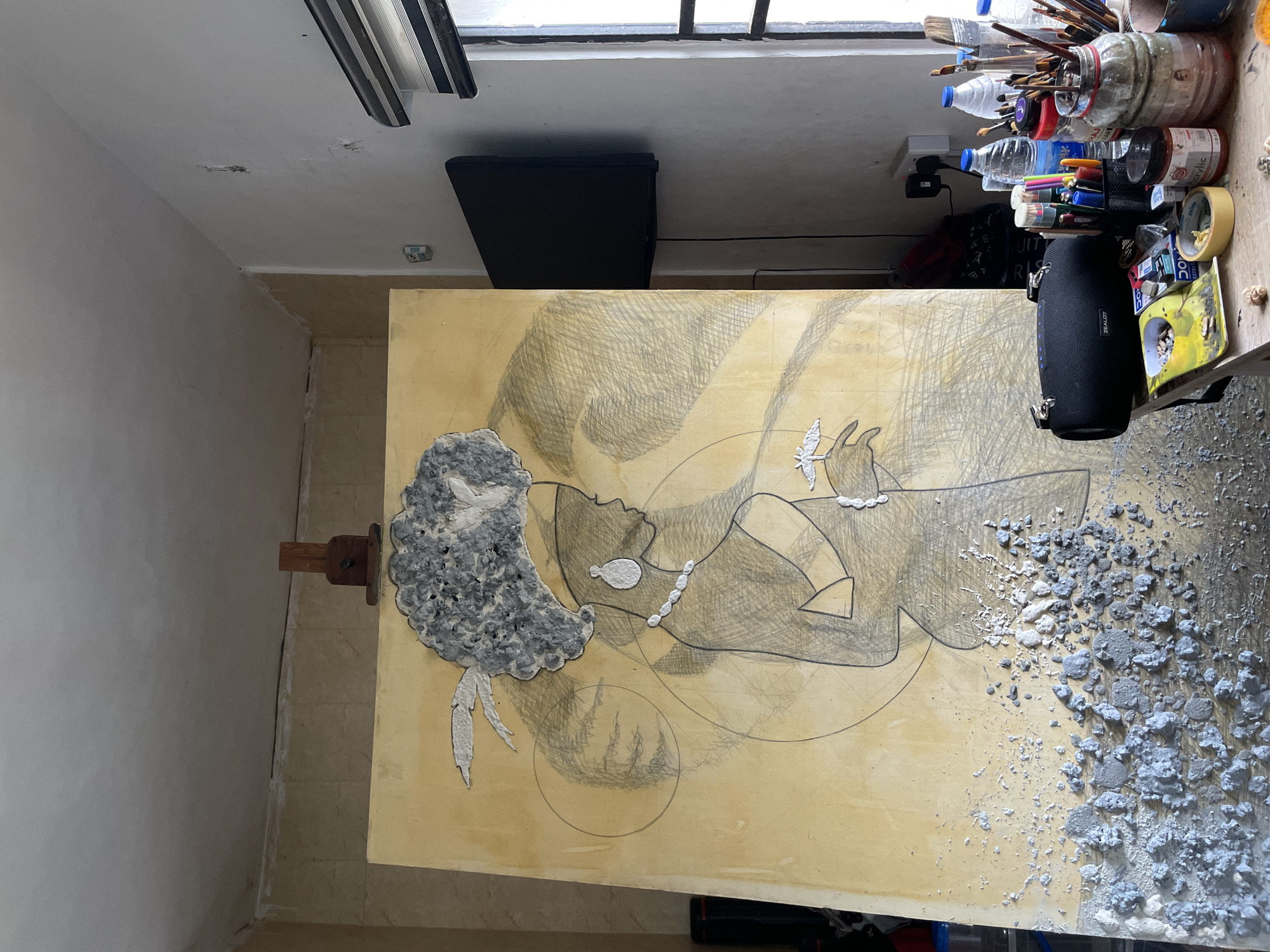 The image shows a room in Ado, Nigeria, with an unfinished artwork. The artwork, propped on an easel, depicts a reclining figure in pencil on a yellow-toned canvas. The figure has an afro-like hairstyle made of grey, plaster-like material. The lower right corner of the canvas is covered with the same grey material, appearing scattered. The room has light yellow walls, a window at the top, and contains objects like paintbrushes, jars, water bottles, and a black speaker on a shelf or table near the window. A black object is also positioned behind the easel, on the ground. The scene suggests an artist's studio.