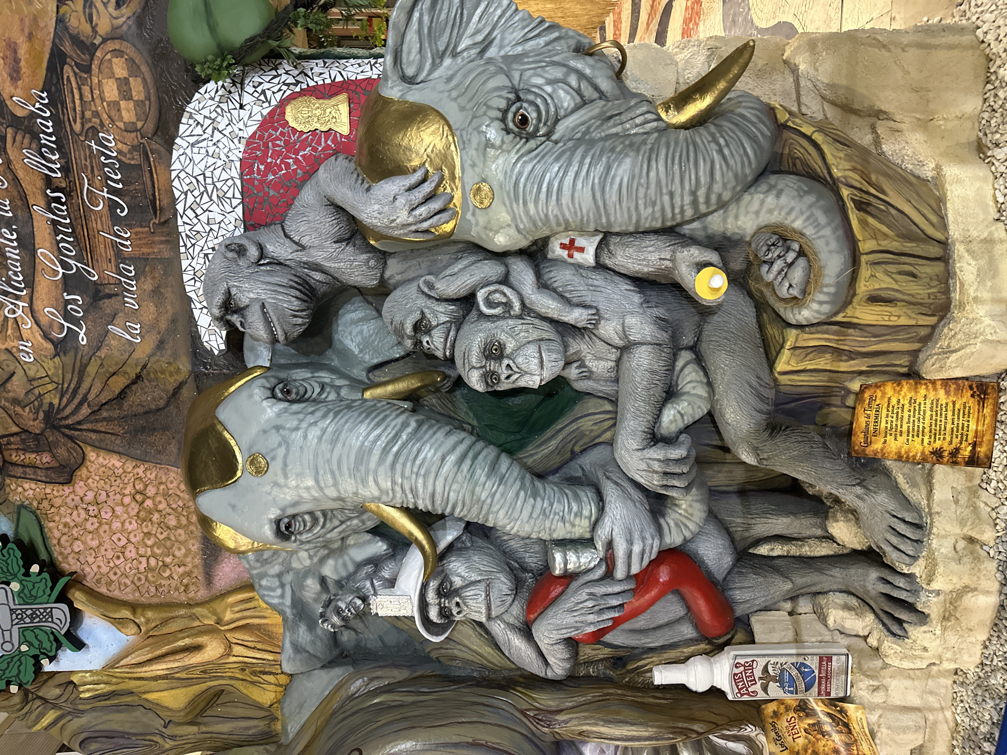 The image depicts a large, elaborate sculpture in Alicante, Spain, featuring a group of gray, realistic-looking chimpanzees and two elephants. The sculpture is part of a larger mural or installation that includes painted elements and text.
In the foreground, several chimpanzees are arranged in a cluster. One chimpanzee, wearing a white hat and holding a red, curved object and a metallic cup, sits prominently. Another chimpanzee is positioned above and behind him, and a third is nestled on the elephant's trunk. A smaller infant chimpanzee is depicted curled up on the elephant's tail. A red cross emblem is visible on one of the chimpanzee's arms.
Two elephants are integrated into the composition. The head of one elephant is to the right, with a golden headdress. Another elephant's trunk and part of its head are on the left. Both elephants have golden accents on their heads and tusks.
Scattered around the base of the sculpture are several objects. A white bottle with a label reading "ANIS TENIS" and "DESDE 1888" is on the left. To the right, a yellow sign with text in Spanish, titled "Guardianes del Tiempo ENFERMERIA," is visible. Another yellow, folded document with similar text is positioned to the left of the "ANIS TENIS" bottle.
The background of the image includes painted imagery and text. White and red mosaic tiles are present behind some of the figures. A portion of a painted mural depicts figures and various patterns. Text in white cursive script reads, "en Alicante, la Los Gorilas llenaba la vida de Fiesta." (In Alicante, the Gorillas filled life with Fiesta.)
The overall environment appears to be an indoor setting, possibly a museum or a public display area, with a floor covered in small white pebbles. The lighting suggests an artificial light source. The sculpture's style is detailed and somewhat whimsical, blending realistic animal forms with fantastical elements.