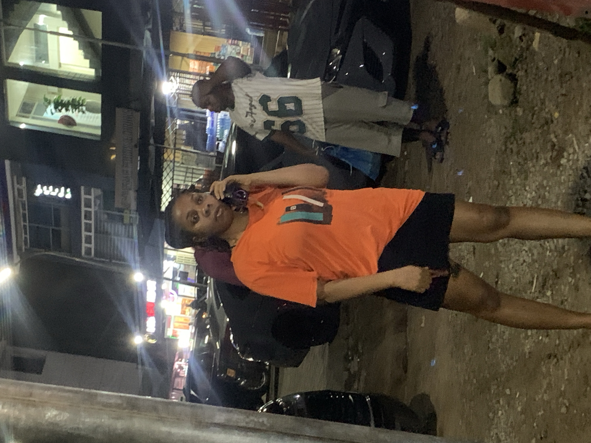 A medium shot captures a Black woman standing outside at night in what appears to be Galadimawa Village, Nigeria. She is facing the camera, holding a dark-colored mobile phone to her right ear with her right hand. The woman is wearing an orange t-shirt with a graphic design on the chest and a black skirt. Her hair is dark and braided, and she has a serious expression. To her right, a Black man stands with his left hand touching his head, wearing a white t-shirt with "56" and "Angel" printed on it, and light-colored shorts. Behind them, multiple dark-colored cars are parked, and a building with illuminated windows and signs is visible in the background. The ground is covered in loose gravel and dirt. The lighting suggests artificial illumination from streetlights or building lights.