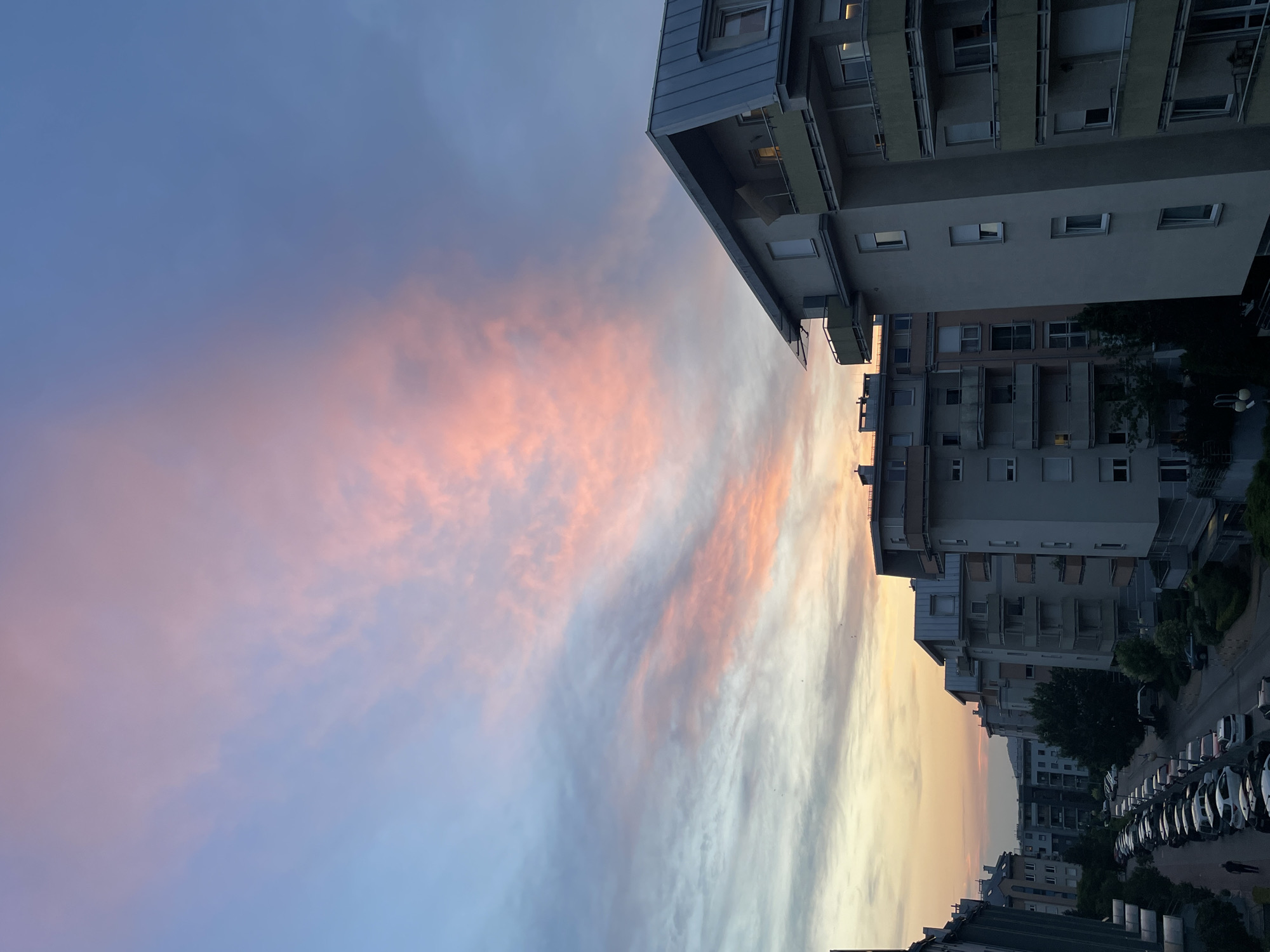This media file depicts a city scene during sunset. The primary subjects are apartment buildings and a sky filled with clouds tinged with pink and orange hues, indicating the time of day is sunset. The scene is a street lined with parked cars, suggesting an urban or suburban residential area.
The activity taking place is implied to be the end of the day, with the buildings illuminated by the setting sun and some windows showing interior lights. A single person is visible walking on the sidewalk. The weather appears clear, with the clouds being a natural phenomenon of the sunset. The location context is unknown, but the architecture suggests a European setting. The overall emotion evoked by the image is peaceful and serene, due to the beautiful colors of the sky. There is no visible text in the image.