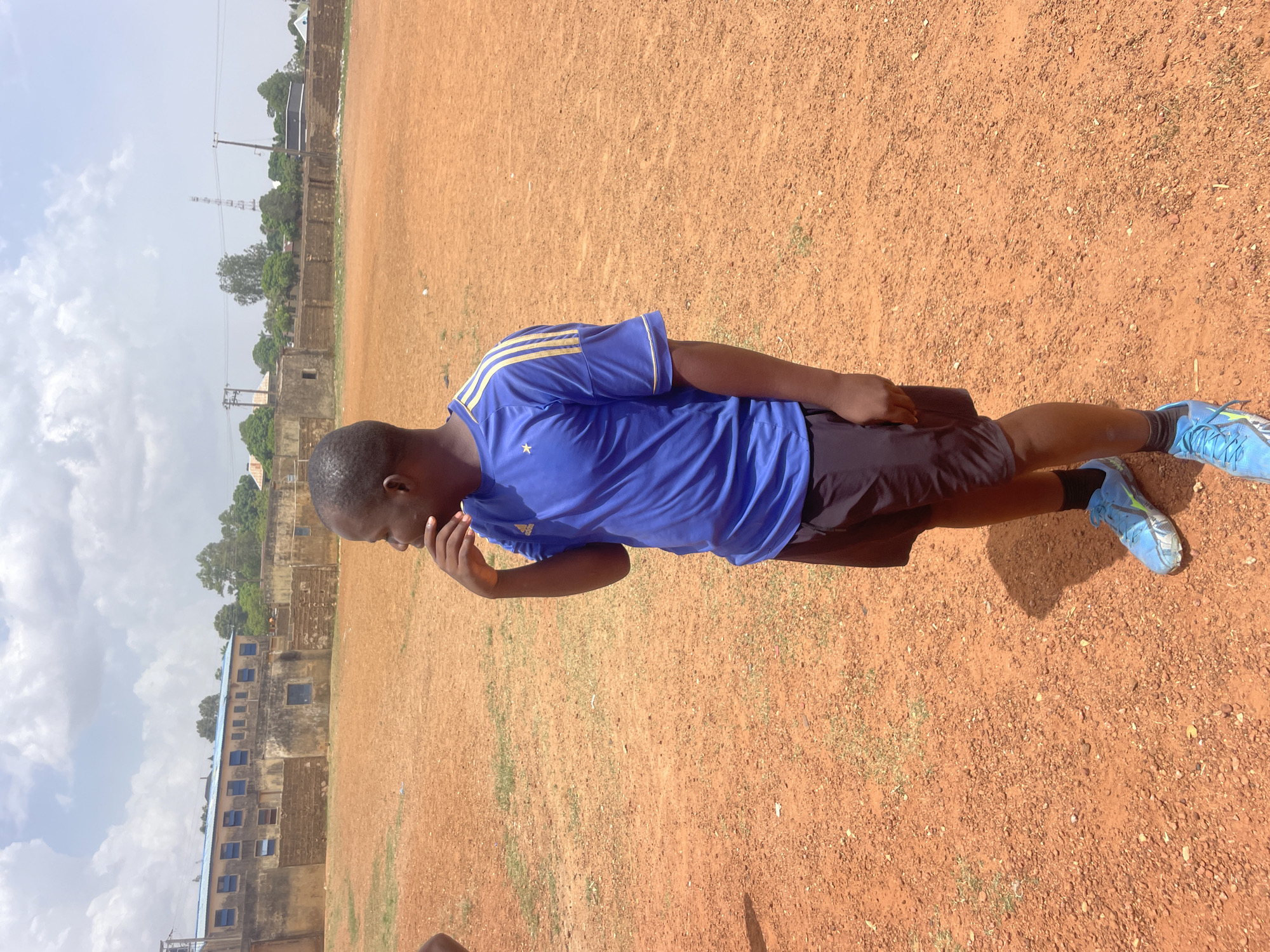 The image shows a young man in athletic attire on a dirt field. He is wearing a blue soccer jersey with yellow stripes, black shorts, and blue soccer cleats. He has his hand to his face in a contemplative gesture. The field has sparse patches of grass. In the background are buildings and a transmission tower. The scene is located in Jos, Nigeria.