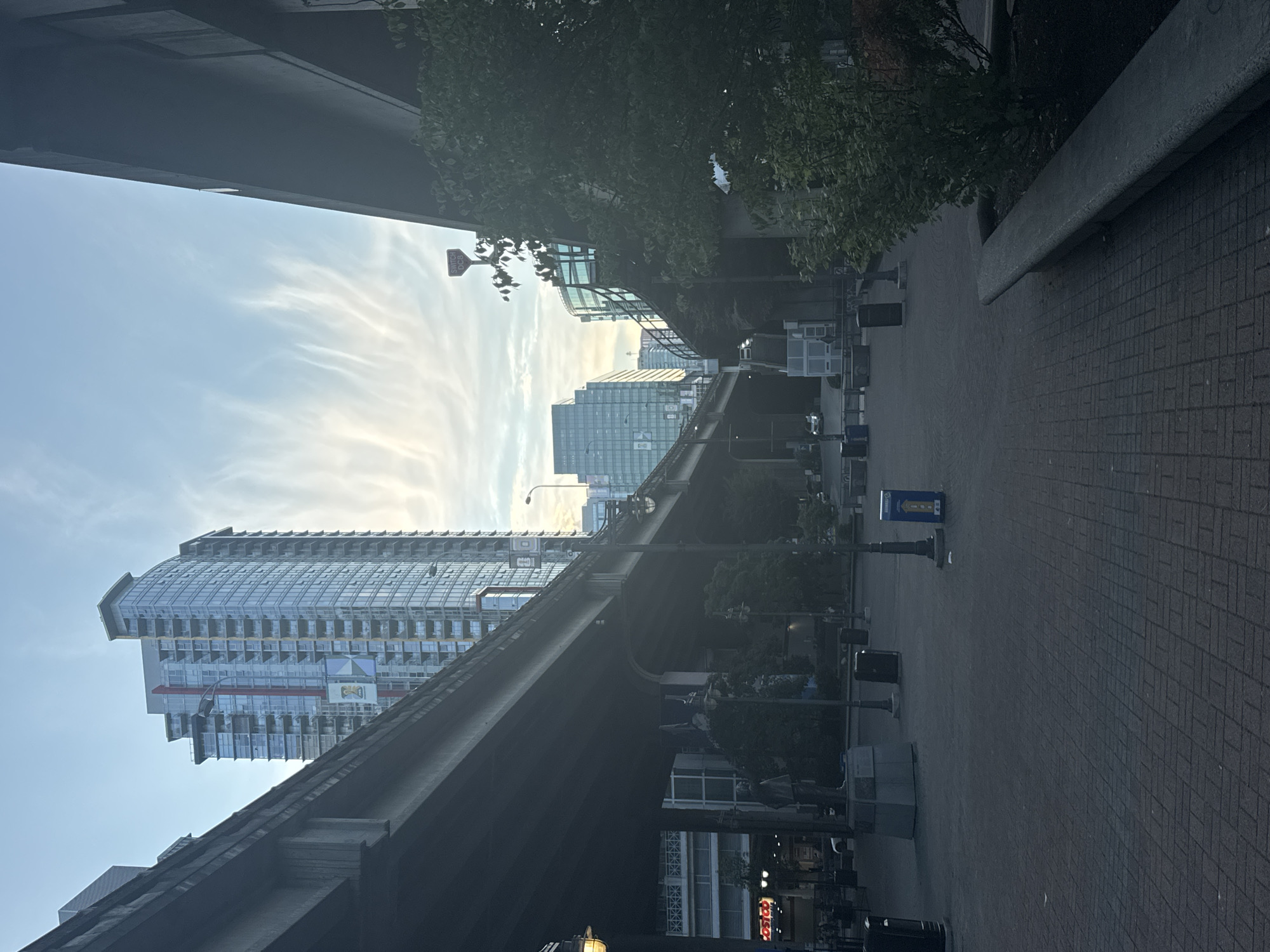 The image captures a street-level view of a city plaza in Vancouver, Canada, featuring a prominent overpass system and modern high-rise buildings. The sky is partly cloudy with wispy cirrus clouds illuminated by the setting sun. In the foreground, a paved plaza with a brick-like pattern is visible, leading towards a large concrete overpass that dominates the left and center of the frame. Several trash receptacles are positioned throughout the plaza. To the right, a dense green bush is situated in a planter box, next to a low concrete seating area. In the background, a tall, slender skyscraper with a glass facade stands out, adorned with banners. Other buildings of varying heights and architectural styles are also visible, suggesting a commercial or residential urban setting. A street lamp is positioned on the right side of the plaza. The overall atmosphere is calm and somewhat muted, possibly due to the time of day and the architectural elements casting shadows.