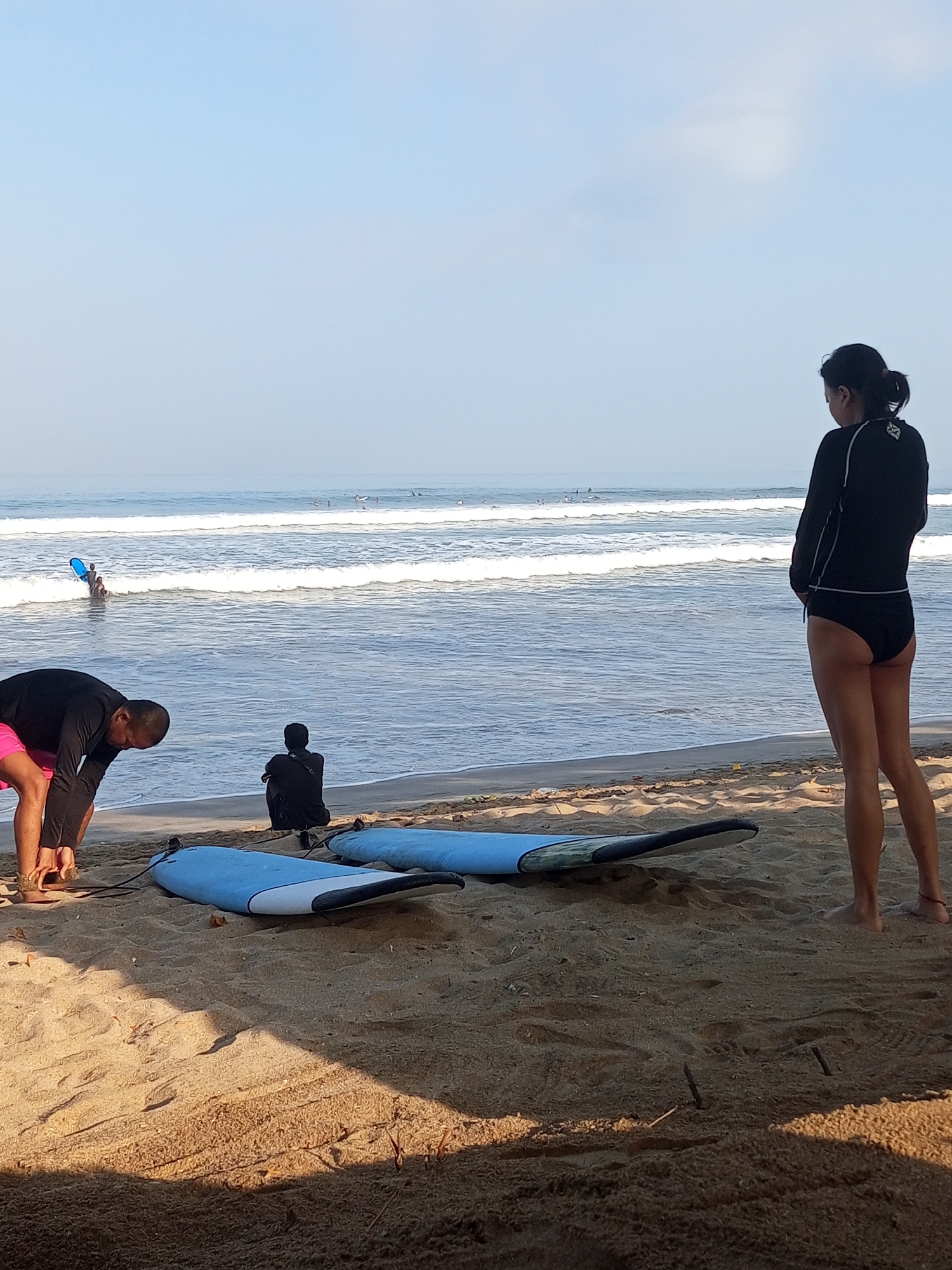 A group of people are gathered on the sandy shore of Kuta beach, Indonesia, with surfboards laid out on the sand. A woman in a black rash guard and bikini bottoms stands facing the ocean, looking out at the waves. A man in a black long-sleeved shirt and pink shorts is bending down, seemingly adjusting his footwear. In the background, several surfers are in the water, riding waves. Another person is seated on the sand, facing away from the camera. The sky is clear and blue, with calm ocean waves rolling onto the shore.