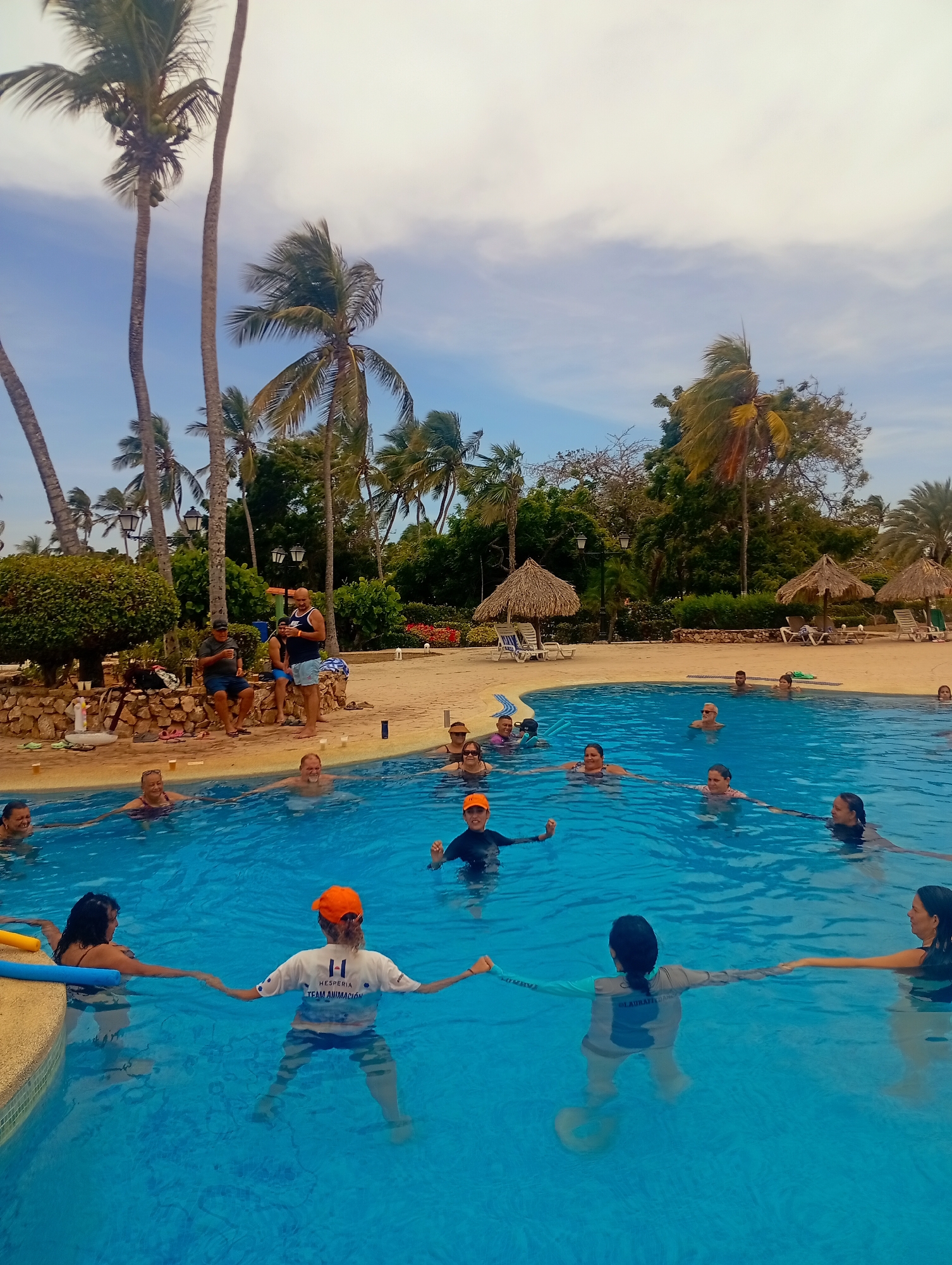 The image shows a group of people participating in a water aerobics class in a swimming pool. The pool is surrounded by a sandy area with palm trees and thatched-roof structures. Several individuals are seated or standing along the poolside, observing the class. Two instructors wearing matching gray shirts and caps, lead the class. Other participants are engaged in exercises within the pool. The sky is partly cloudy. This location is in Playa El Agua, Venezuela.
