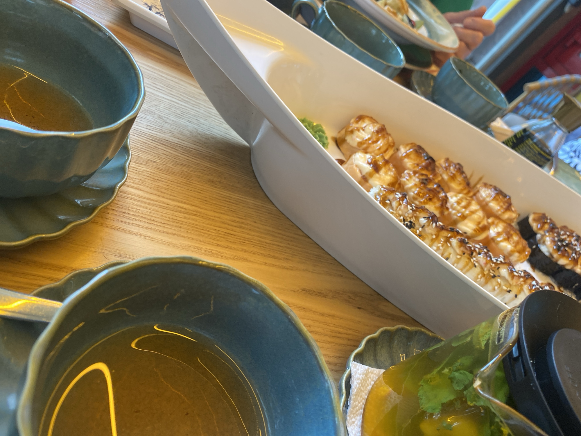 The image depicts a close-up, angled view of a table setting at what appears to be a Japanese restaurant. In the foreground, two teal-colored ceramic bowls are visible, each containing a golden-amber liquid, likely tea or broth. To the left, a white serving dish holds several pieces of grilled sushi, topped with a glossy sauce and sesame seeds. Behind the sushi, a glass teapot filled with green liquid and mint leaves is positioned on a dark green scalloped saucer, suggesting a hot beverage is being served. The table surface is a light-colored wood with a visible grain. In the background, a hand is reaching for something, and part of a bottle with a dark label is also visible. The overall atmosphere suggests a meal in progress, with a focus on Japanese cuisine and beverages. There is no indication of the specific location within the city of Tuymazy, Russia, or any other geographical markers.