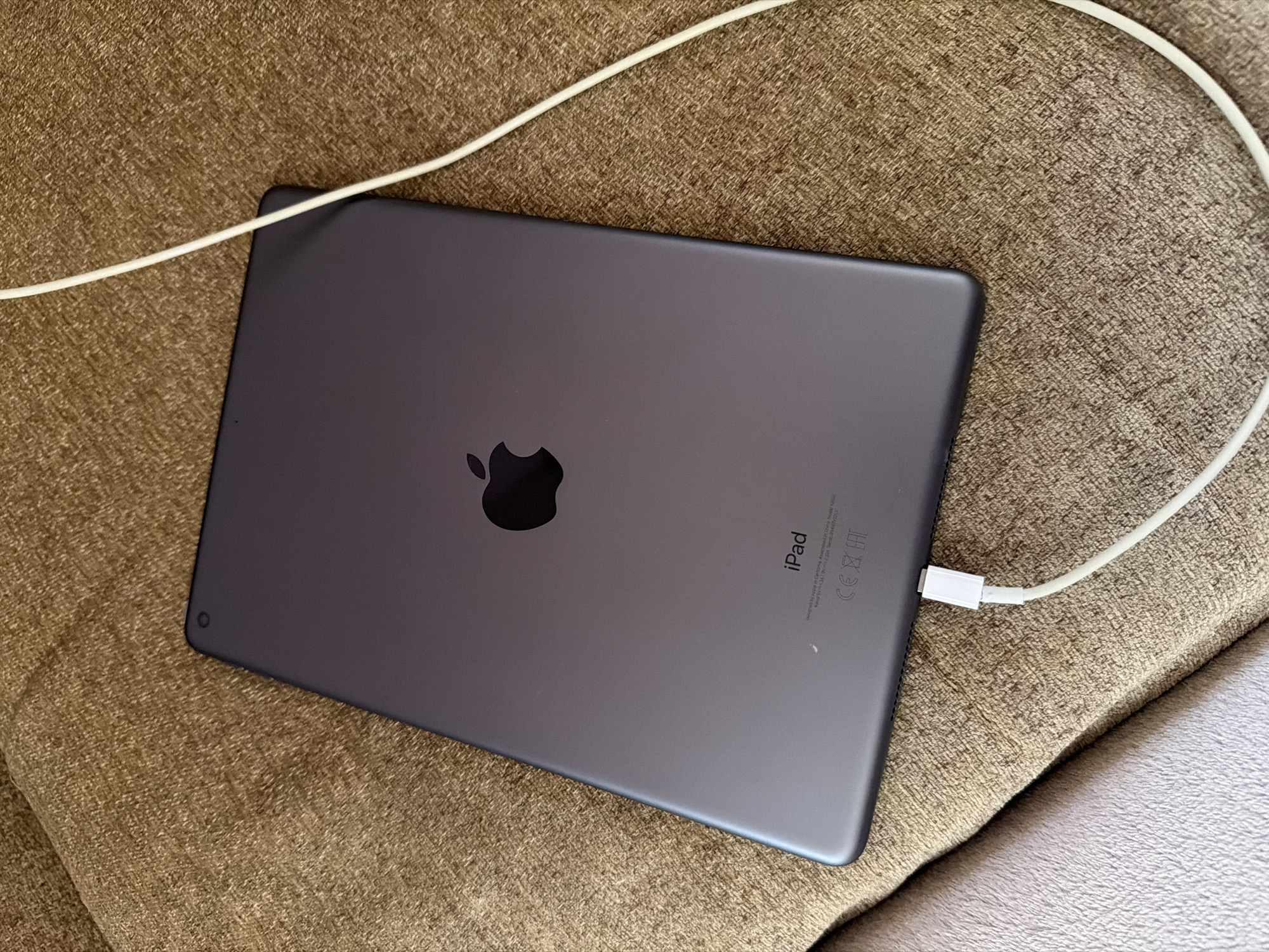The image shows the back of a dark gray Apple iPad placed on a textured brown fabric surface, likely a couch or chair. A white charging cable is connected to the iPad and extends out of the frame. The Apple logo is prominently displayed in the center of the iPad's back. Below the logo, the word "iPad" is printed, followed by smaller text indicating manufacturing details and certifications, including "Designed by Apple in California," "Assembled in China," and the CE and FCC marks. The scene is an indoor setting, with no clear indications of time of day or weather. The overall impression is one of a device ready for charging or in the process of being charged. The fabric surface has a visible woven texture, and the lighting suggests a moderately lit indoor environment. There are no people visible in the image.