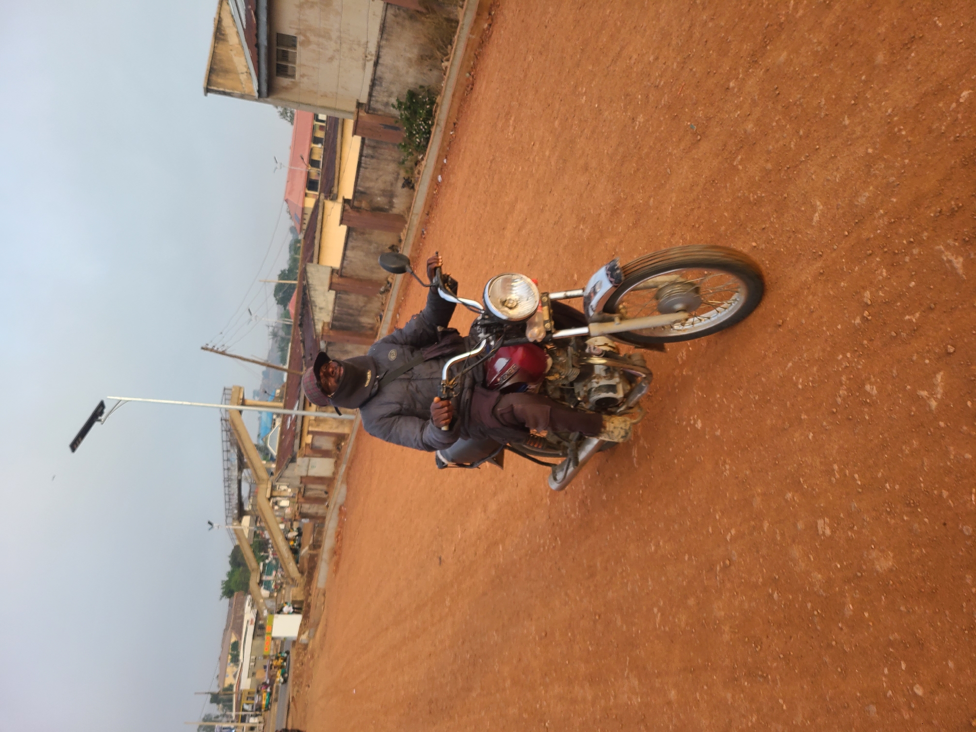 A man wearing a jacket and head covering is riding a motorcycle on a dirt road in Jos, Nigeria. The road is reddish-brown and appears unpaved. Buildings are visible in the background, with some appearing weathered. A tall, metal structure, possibly a crane, is also visible in the background. A streetlight is present near the side of the road. Other vehicles and objects can be seen further down the road. The sky is overcast.