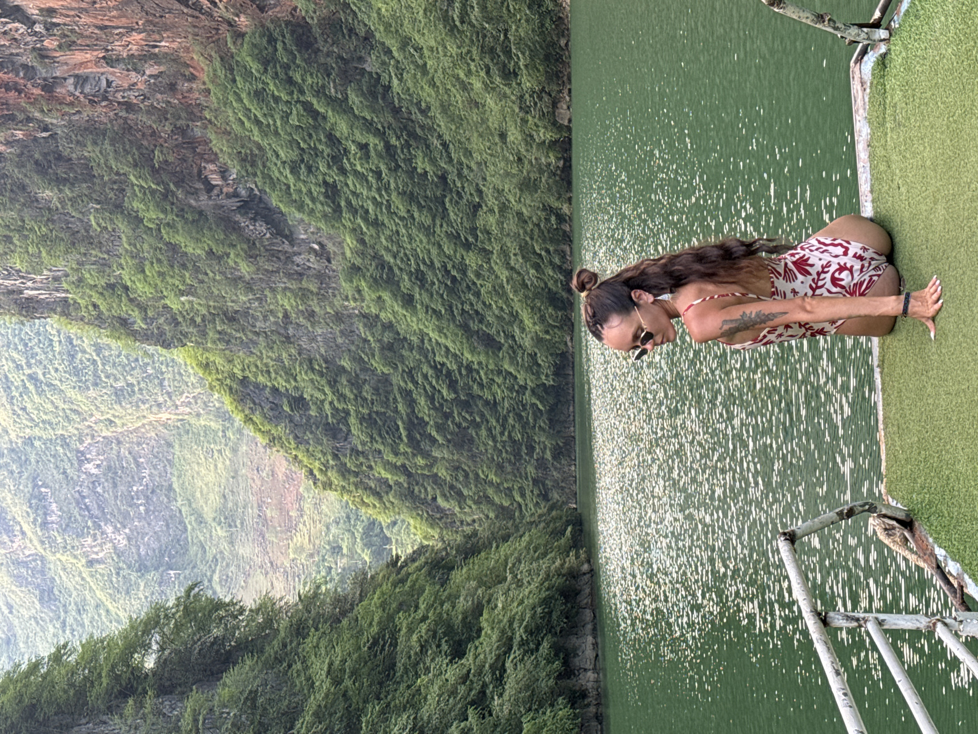 An eye-level shot captures a woman in a patterned swimsuit, kneeling at the edge of a platform adjacent to a body of water. The woman has long brown hair styled in a bun, and she wears sunglasses. A tattoo is visible on her upper arm. The platform has artificial green turf. The water surface glitters with reflected light. Across the water, a steep, verdant hillside dominates the frame. The location is Đồng Văn Commune, Vietnam.