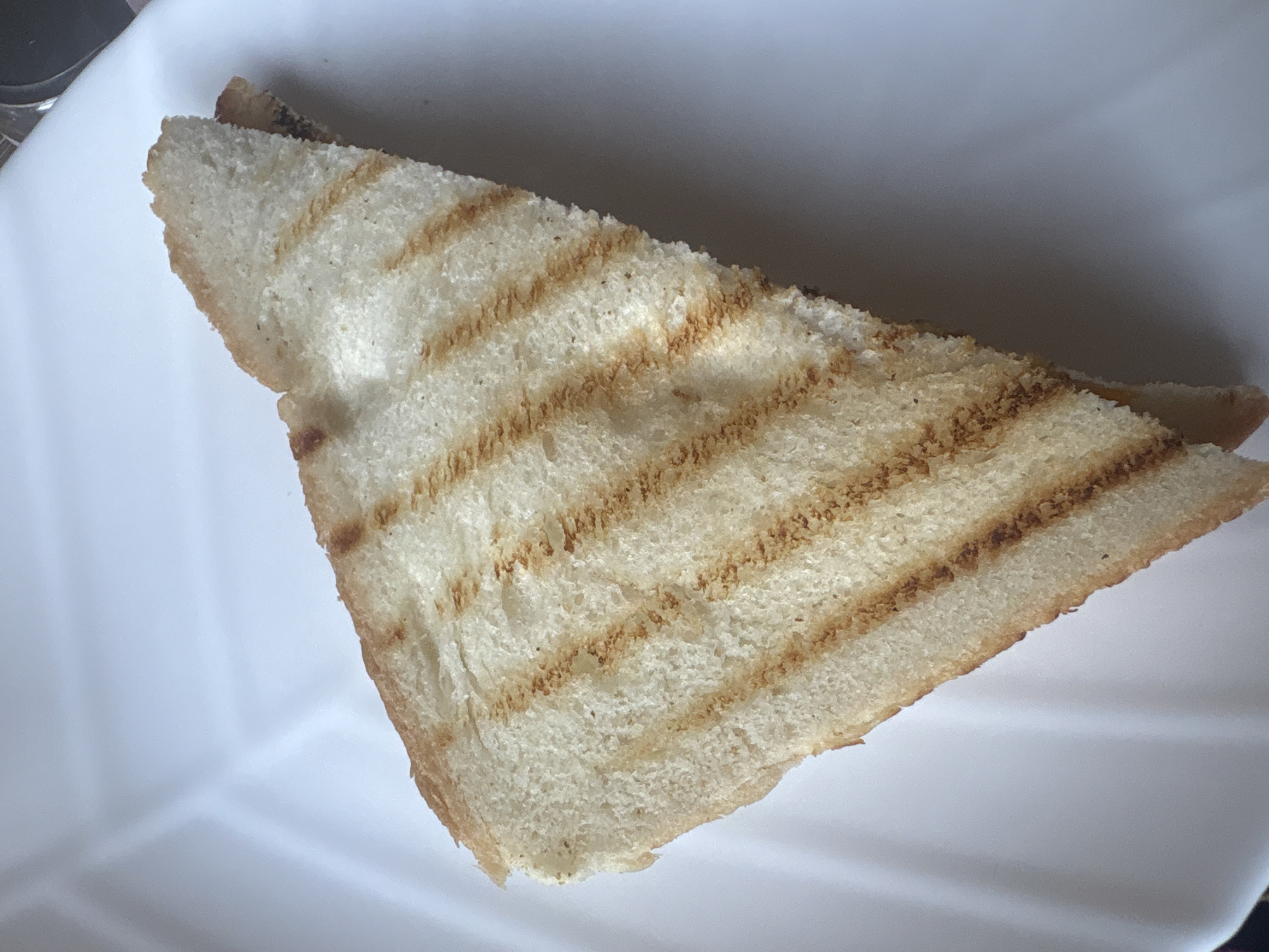 A triangular slice of grilled white bread is placed on a white, segmented plate. The bread has parallel grill marks and a light golden brown color. The plate has a subtle texture and is illuminated from above, casting soft shadows. The background is out of focus and appears to be a neutral, light-colored surface, possibly a tablecloth or countertop. The overall scene suggests a simple meal setting, likely indoors, with no indication of the specific location in Aktobe, Kazakhstan, or any people present.