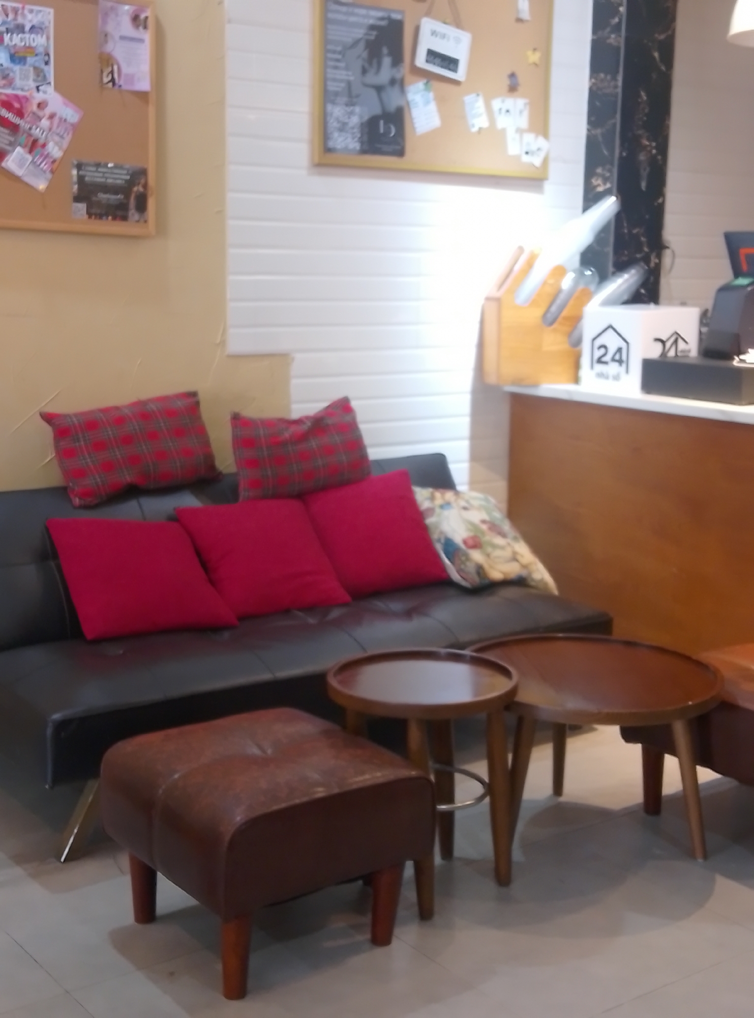 This indoor scene, located in Bac Nha Trang Ward, Vietnam, features a furnished waiting or lounge area. A black faux-leather sofa with metallic legs is positioned on the left, adorned with three solid red throw pillows and two red plaid pillows. A single floral-patterned cushion rests on the sofa's right armrest. In the foreground, a brown leather tufted ottoman with four wooden legs is visible. Two round, dark wooden coffee tables with splayed wooden legs are situated between the sofa and the foreground.
The background consists of a cream-colored wall on the left, a white horizontal paneled wall in the center-right, and a dark marble-patterned vertical column on the far right. Two cork boards are mounted on the walls. The left cork board displays various pinned papers, including one with "KACTOM" and a "WIFI" sign. The right cork board features a framed black and white print of a stylized figure, along with other smaller papers and what appear to be QR codes.
On a light wooden counter to the right, a wooden organizer, possibly a knife block, holds several long, white, pointed objects. Adjacent to this, two white cubic displays are present; one prominently features a black outlined house icon with "24" and Vietnamese text "nhà vỗ". A dark gray point-of-sale terminal is partially visible on the far right of the counter. The area is devoid of people or direct action.