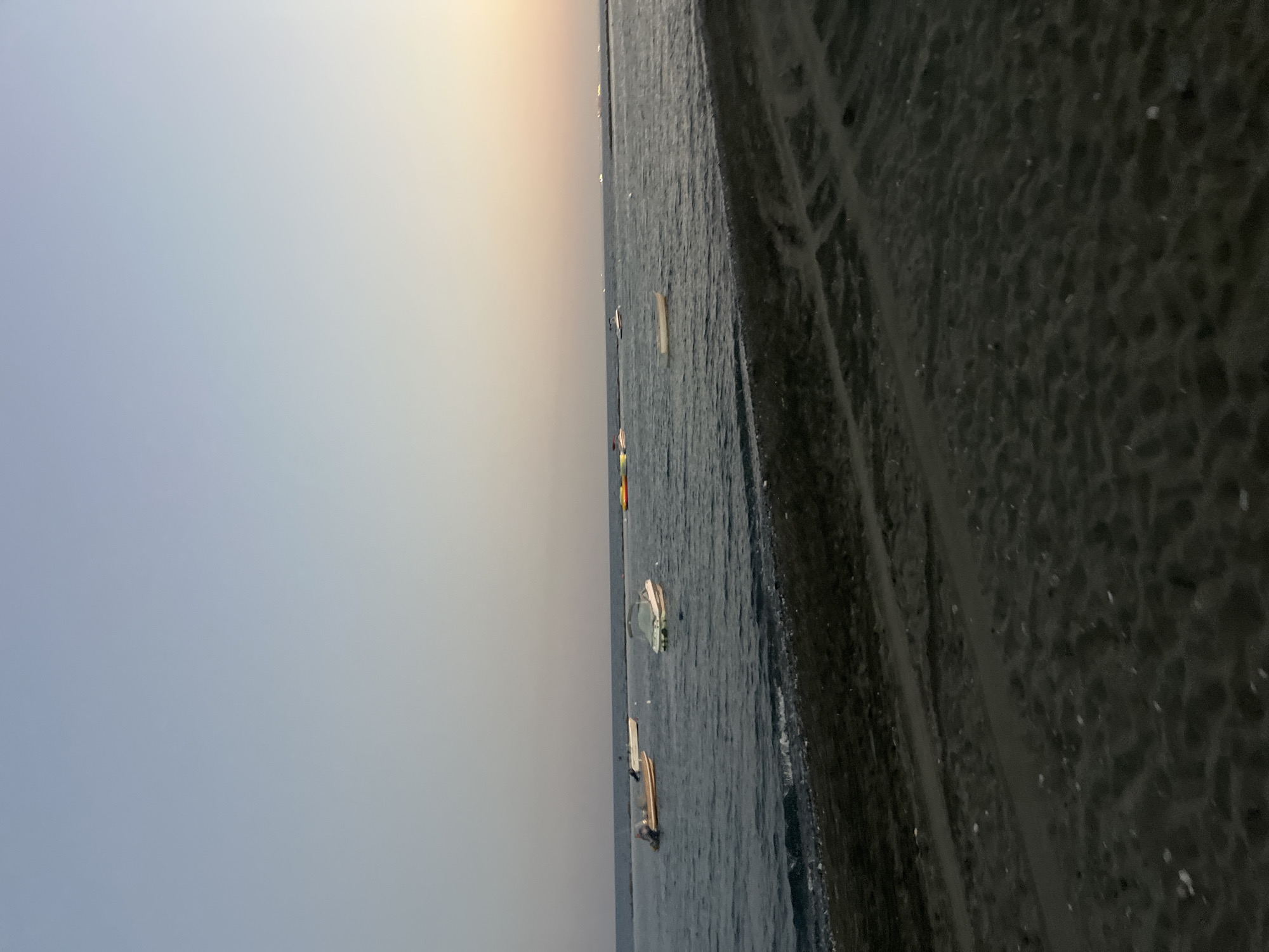 The image depicts a serene coastal scene at dusk in Agios Tychona Community, Cyprus. The sky is a gradient of soft blue and pale yellow, indicating the sun has recently set or is about to rise. The calm sea stretches to the horizon, with several small boats scattered across its surface. In the foreground, a dark, sandy beach is visible, with tire tracks or trails etched into the sand. The overall atmosphere is peaceful and tranquil.