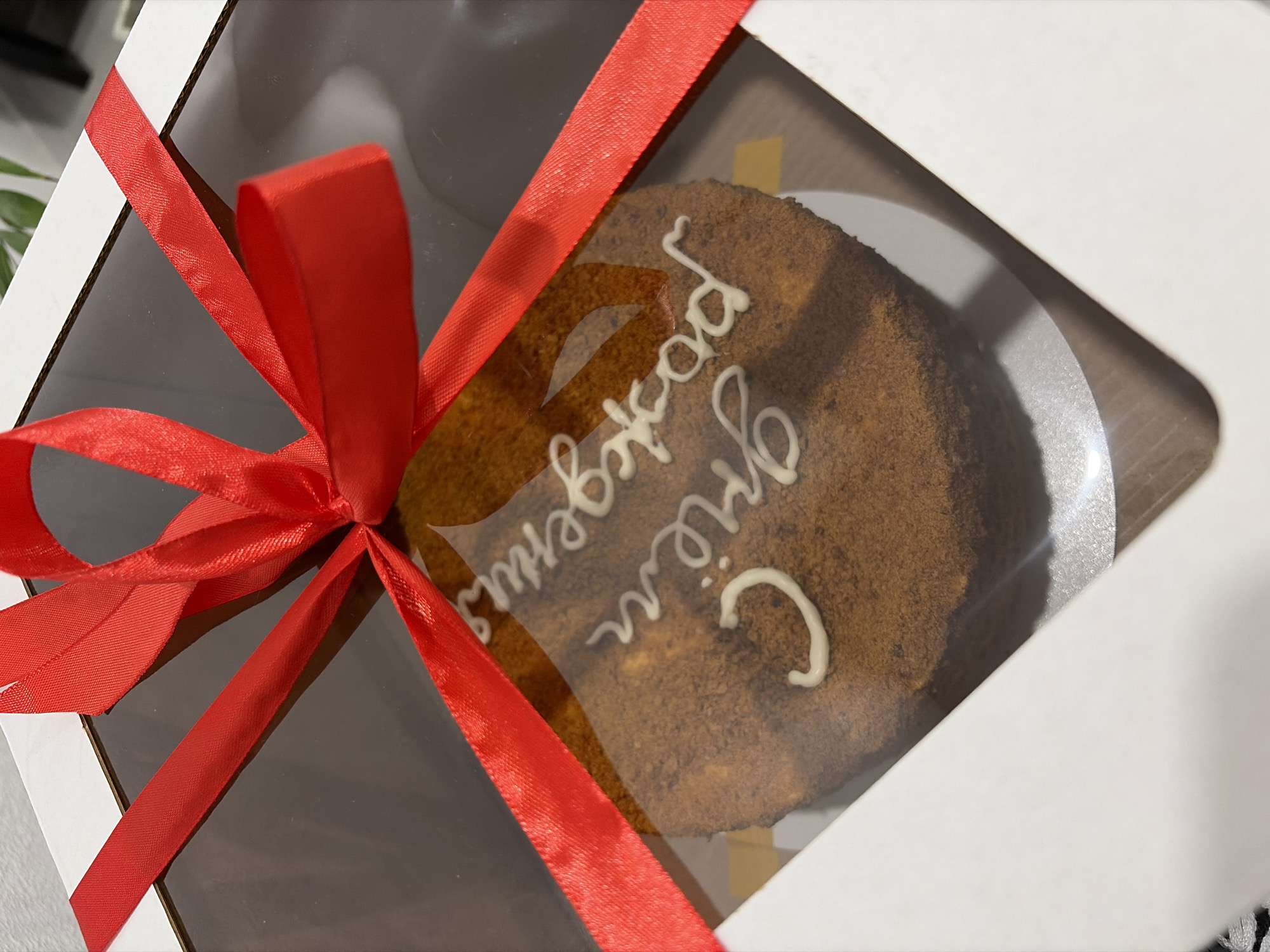 A cake inside a white square box is tied with a red ribbon. The cake appears to be brown with white frosting in a script. The box is sitting on a surface that looks like a floor or table. The frosting reads "С днём рождения" which translates to "Happy Birthday." The box is a see-through box. The exact location in Sarapul, Russia cannot be determined from the image.