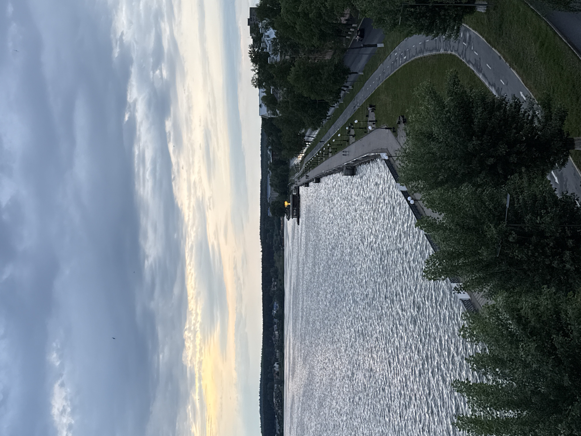 The image depicts a waterfront scene in Votkinsk, Russia. To the left, the sky is partly cloudy, with some sections showing a hint of sunset. A body of water, presumably a lake or river, occupies a significant portion of the frame, displaying light ripples on its surface. A shoreline is visible, lined with trees and some buildings in the distance. A concrete promenade runs parallel to the shoreline, equipped with lampposts along its edge. Adjacent to the promenade, there is a paved path intended for pedestrians or cyclists. A line of trees grows between the promenade and the path.